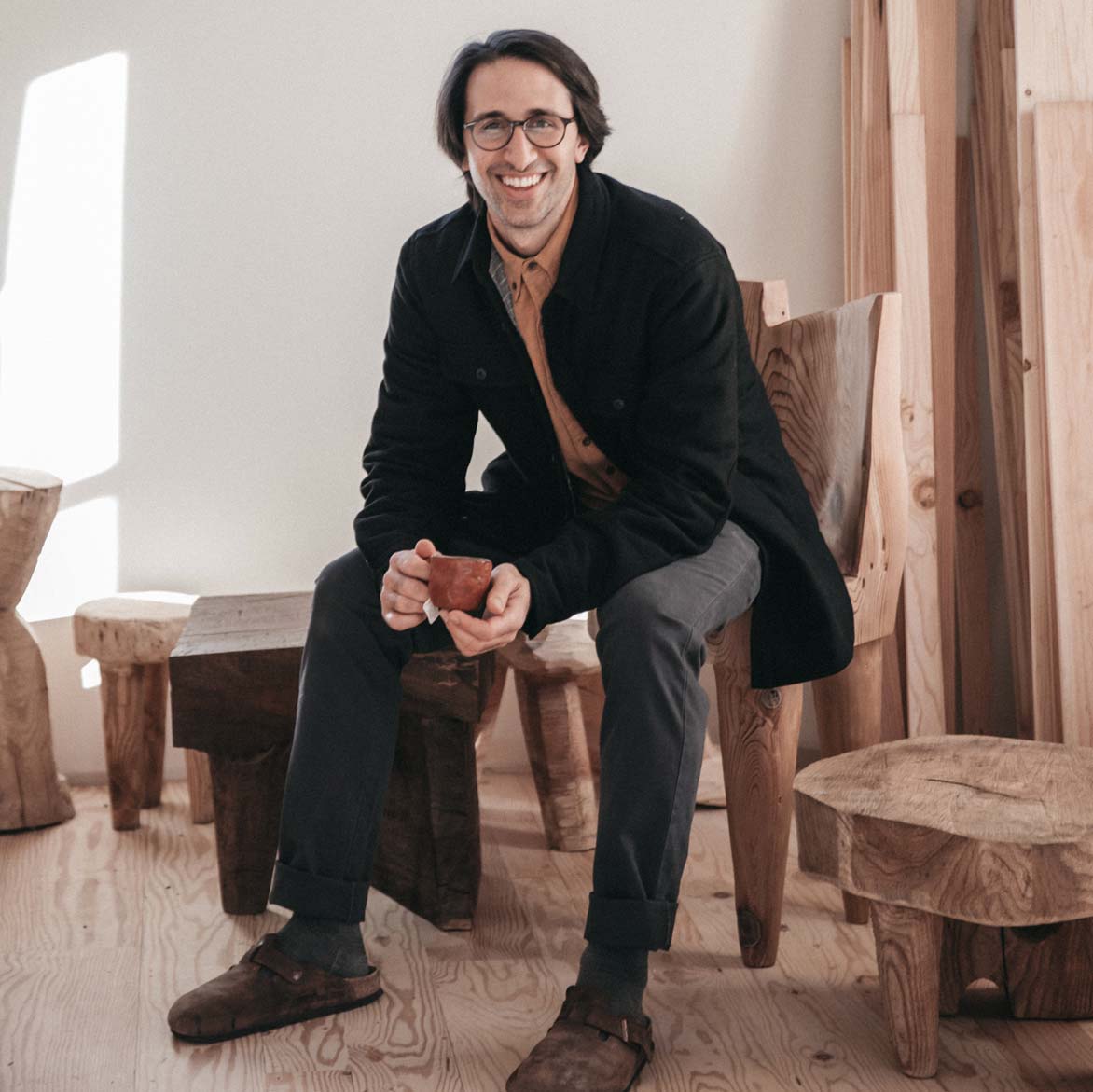 A smiling man with glasses, wearing a black jacket and gray pants, sits on a wooden chair holding a cup. He is surrounded by rustic wooden furniture in a sunlit room with light-colored wooden floors.