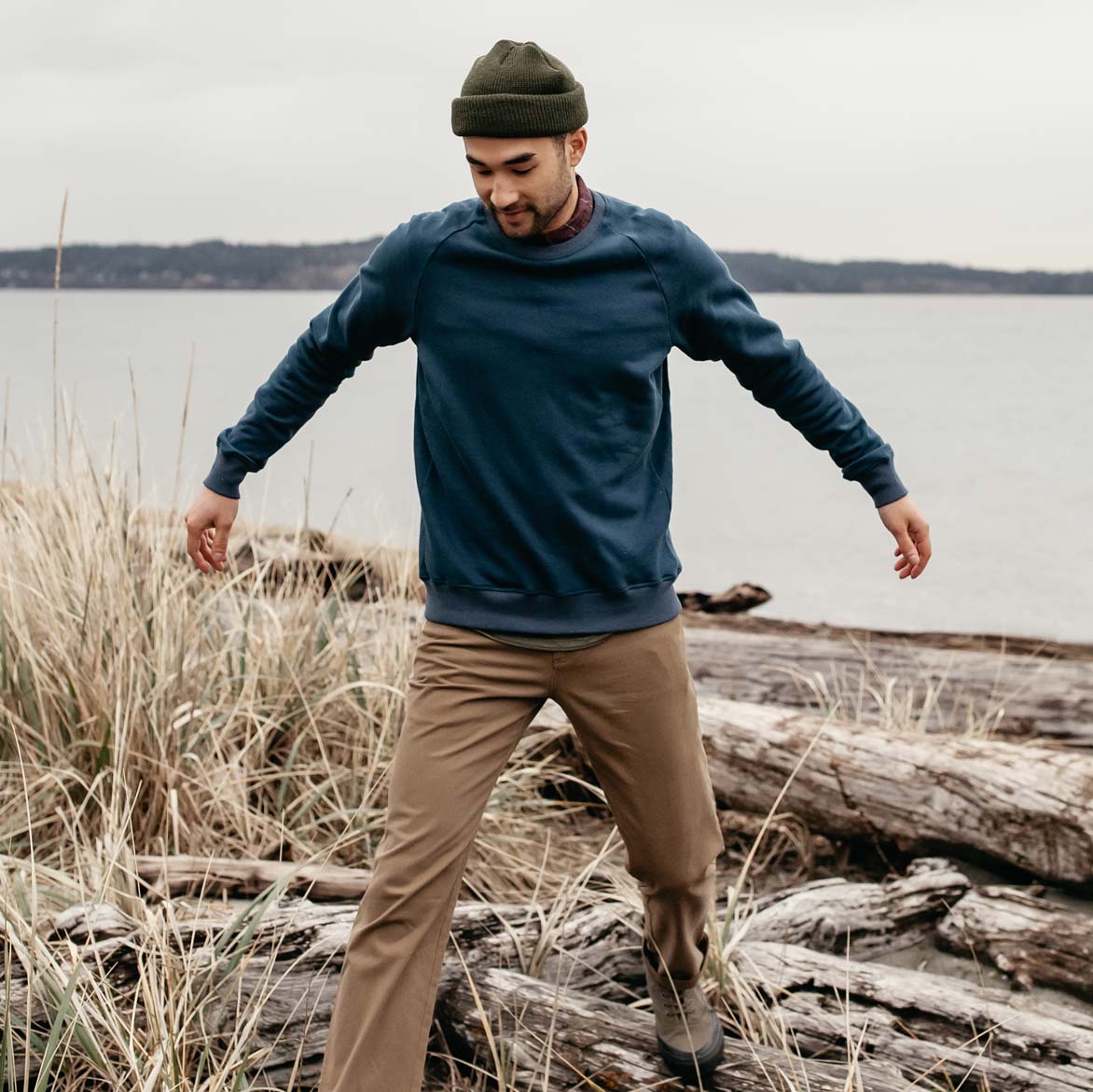 A person wearing a green beanie, blue sweatshirt, and tan pants walks carefully over driftwood and grass by a lakeshore on an overcast day.