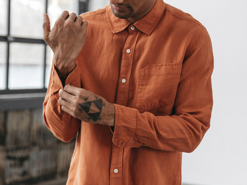 A person wearing an orange button-up shirt fastens the cuff of their sleeve. Their face is not shown, and they have a geometric black tattoo on the back of their hand. A window with black framing is in the background.