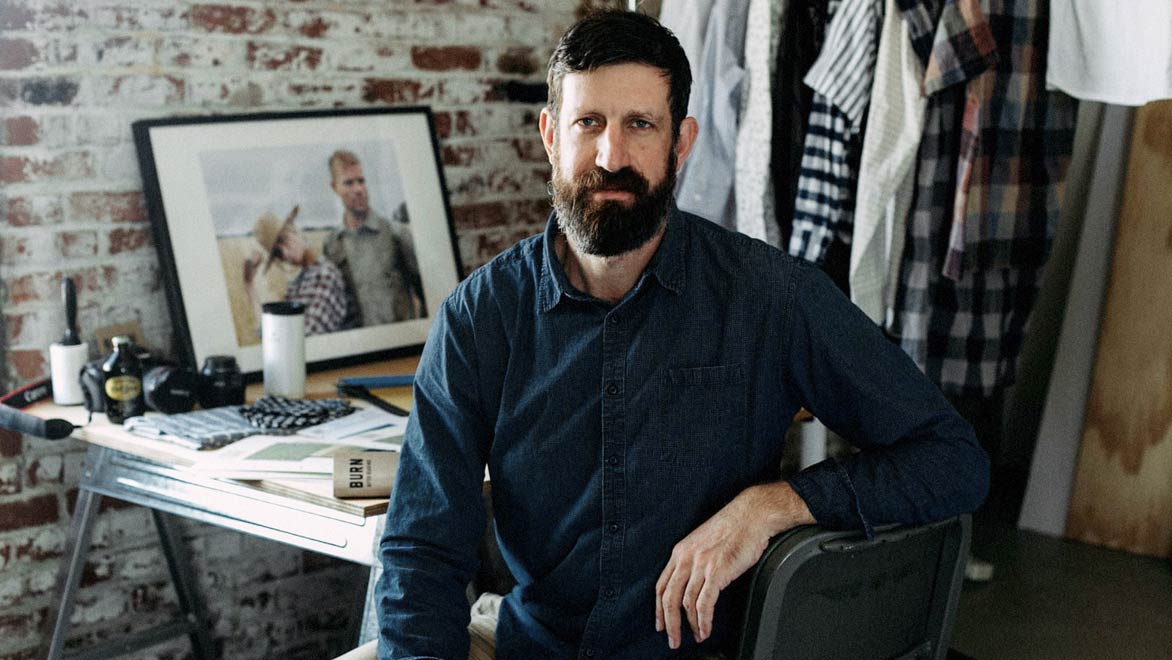 A bearded man in a denim shirt sits on a chair in a studio with a brick wall, clothing hanging, and a cluttered desk with papers, a framed photo, and personal items in the background.