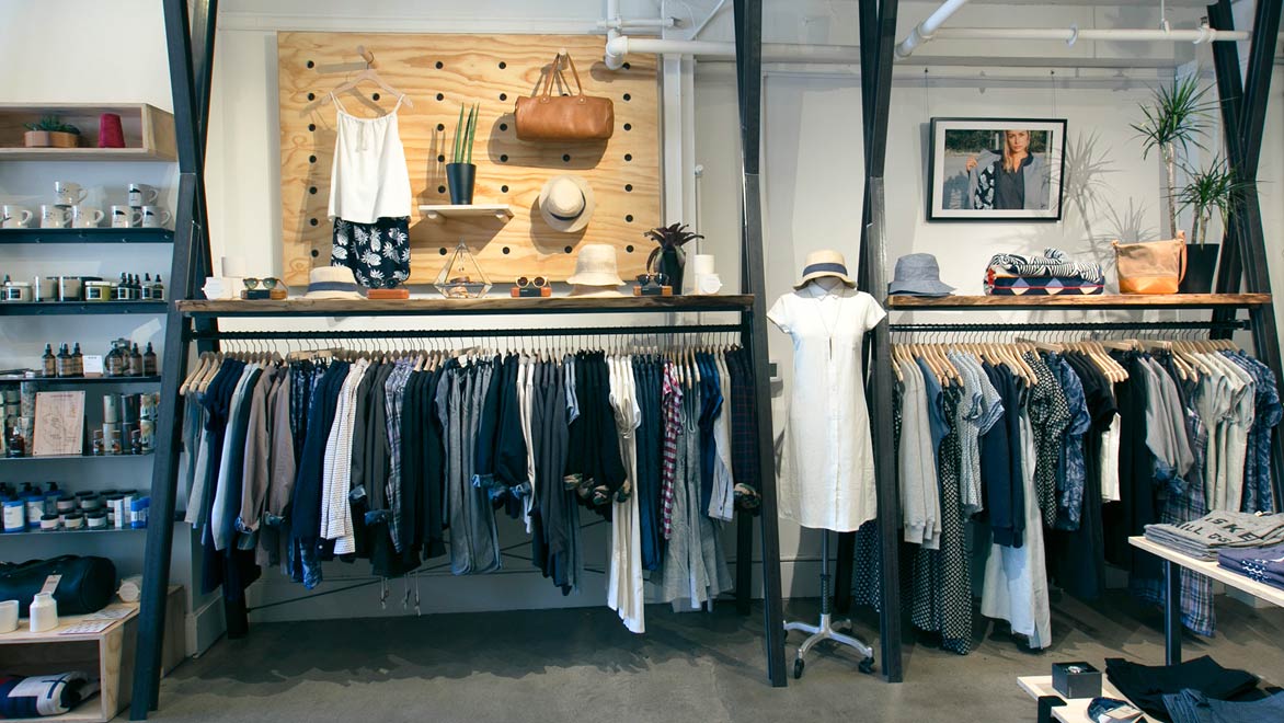A boutique clothing store displays neatly hung shirts, dresses, and pants under a shelf with hats, bags, and sunglasses. The wall has decorative items, a framed photo, and plants. Shelves on the left hold various small goods.