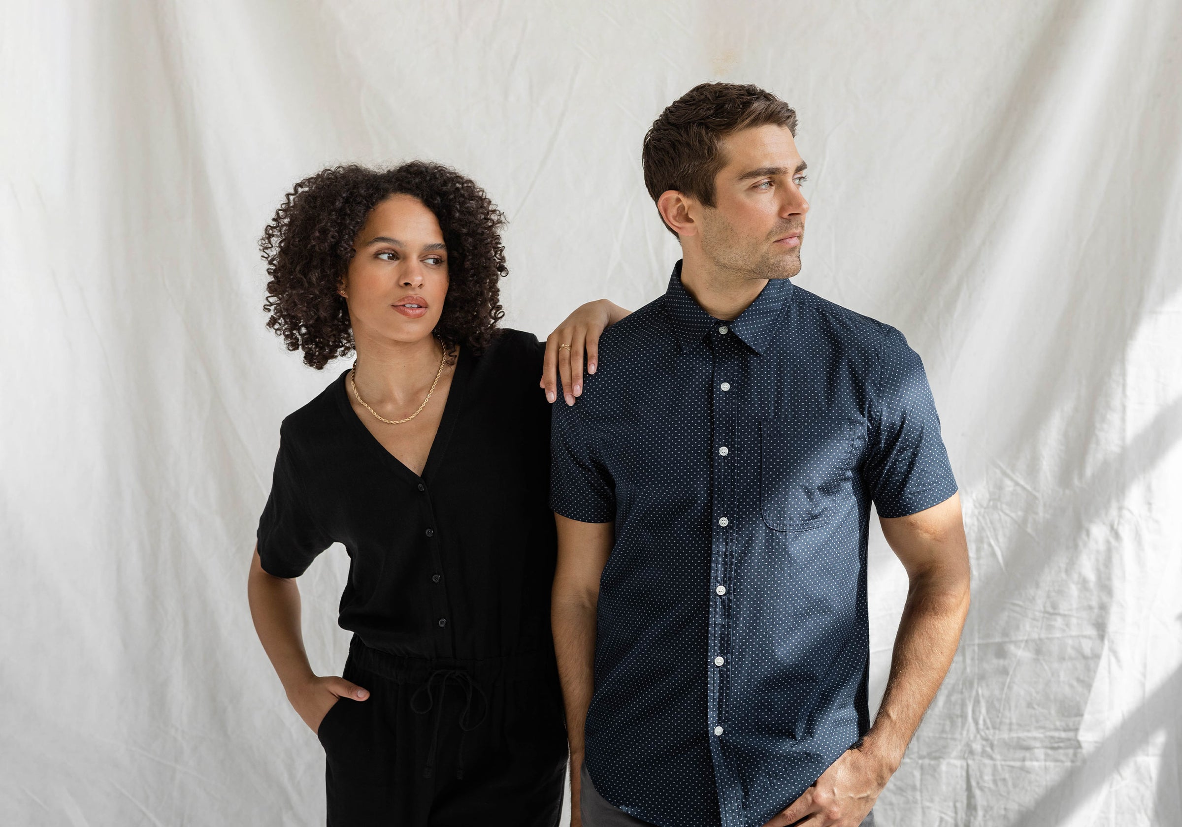 A woman in a black jumpsuit stands with her arm resting on the shoulder of a man in a navy short-sleeve shirt. Both look off to the side against a plain white backdrop.
