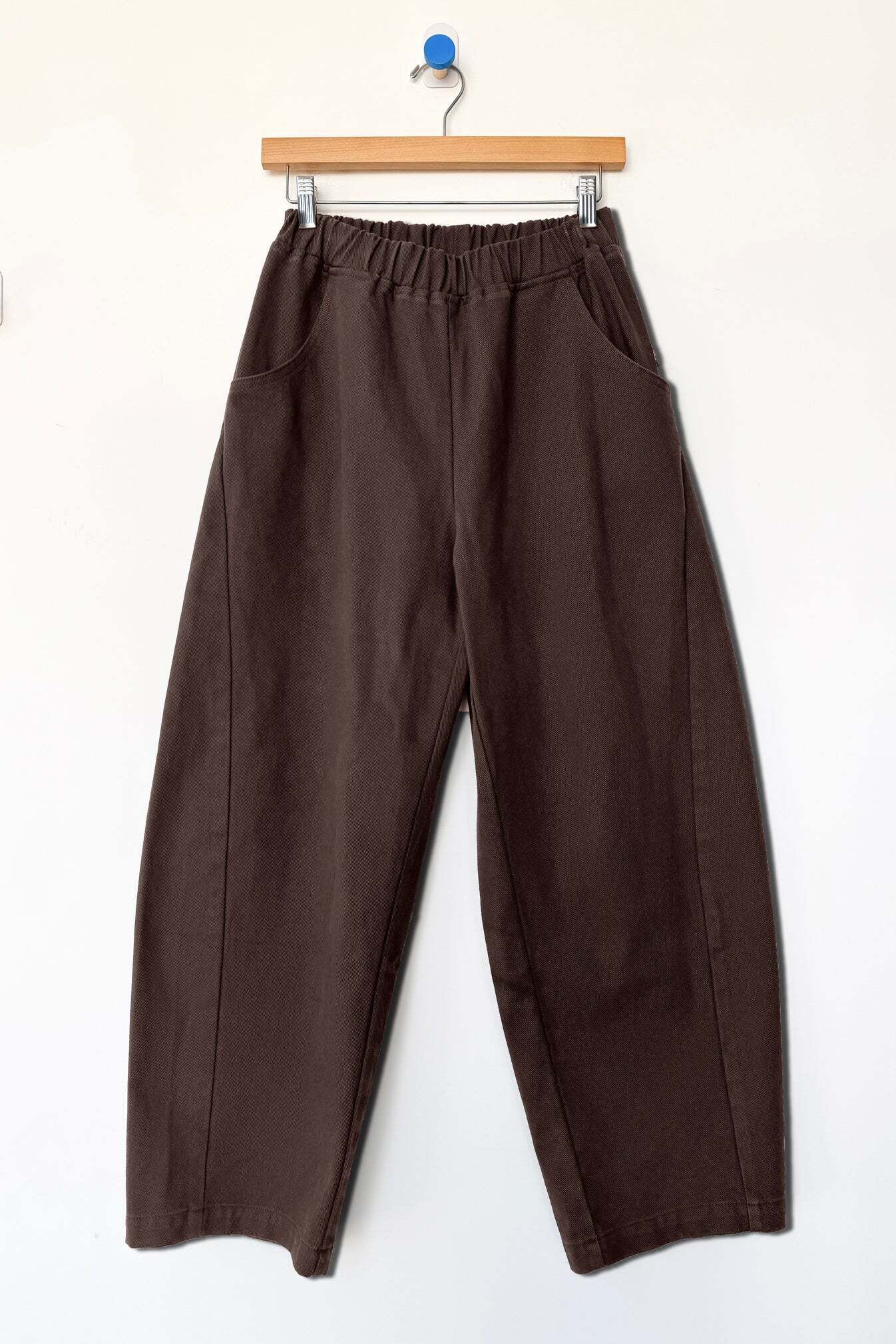 The Le Bon Shoppe - Arc Pant in Umber by Le Bon Shoppe features a loose fit and high-waisted design, crafted from heavyweight denim with an elastic waistband and side pockets. The pants are displayed on a wooden hanger against a plain white wall.