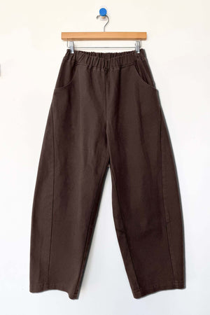 The Le Bon Shoppe - Arc Pant in Umber by Le Bon Shoppe features a loose fit and high-waisted design, crafted from heavyweight denim with an elastic waistband and side pockets. The pants are displayed on a wooden hanger against a plain white wall.