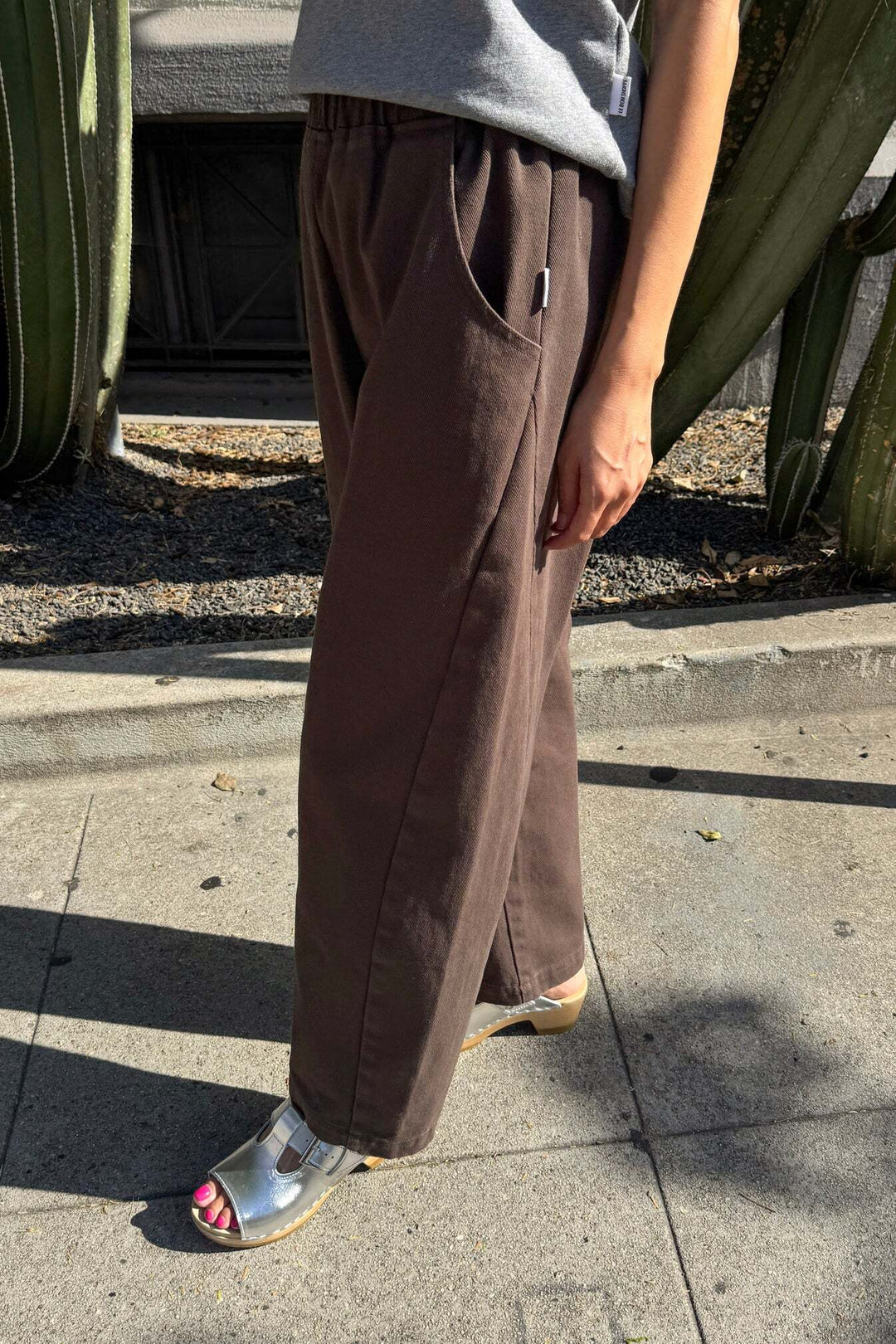 A person wearing Le Bon Shoppe - Arc Pant in Umber stands on a sunny sidewalk near large cacti, paired with a grey top and silver sandals. Only the lower half of their body is visible.