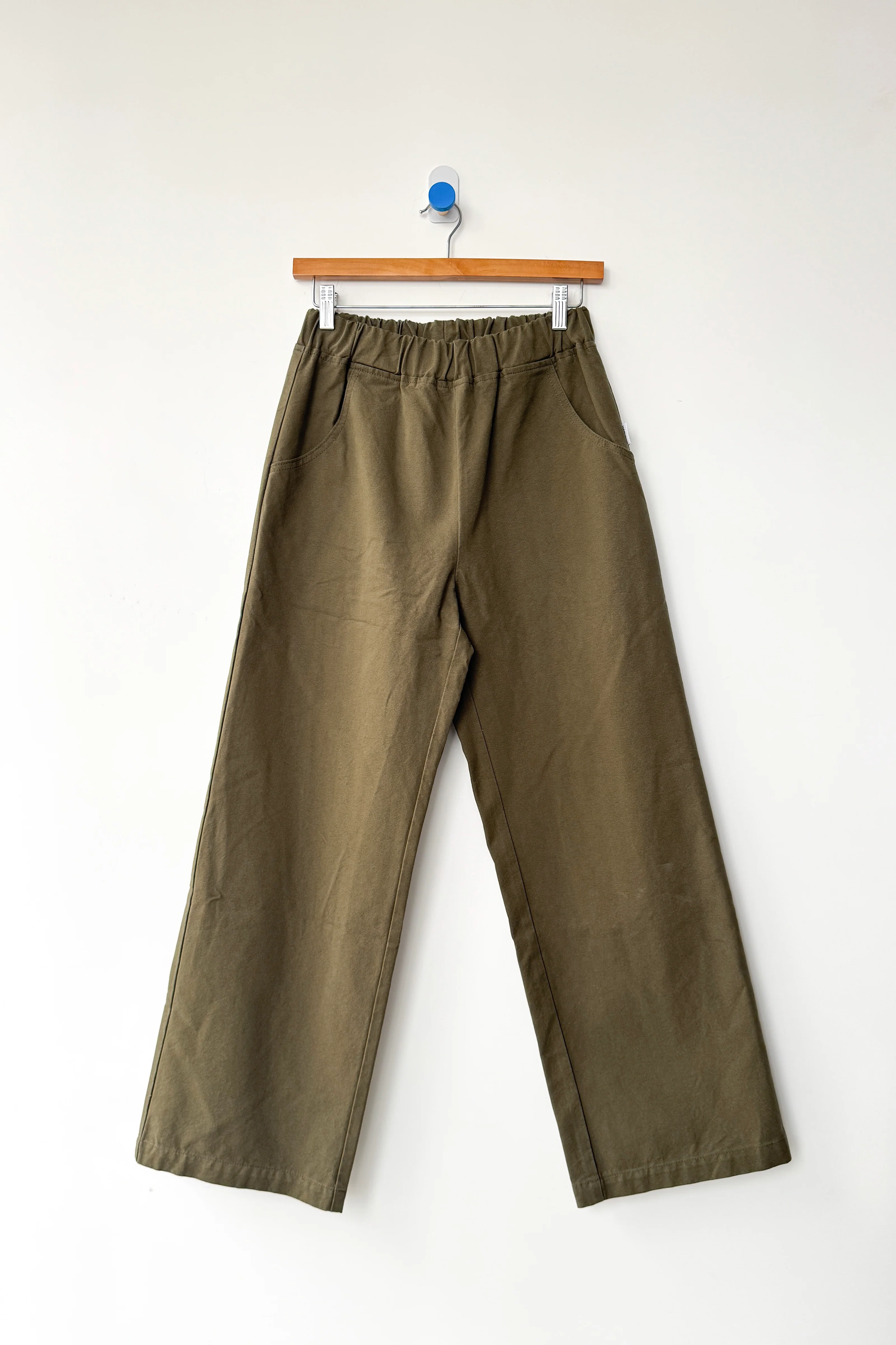 The Le Bon Shoppe Work Pant in olive, a wide-leg cotton canvas style with an elastic waistband and side pockets, hangs on a wooden hanger against a plain white wall.