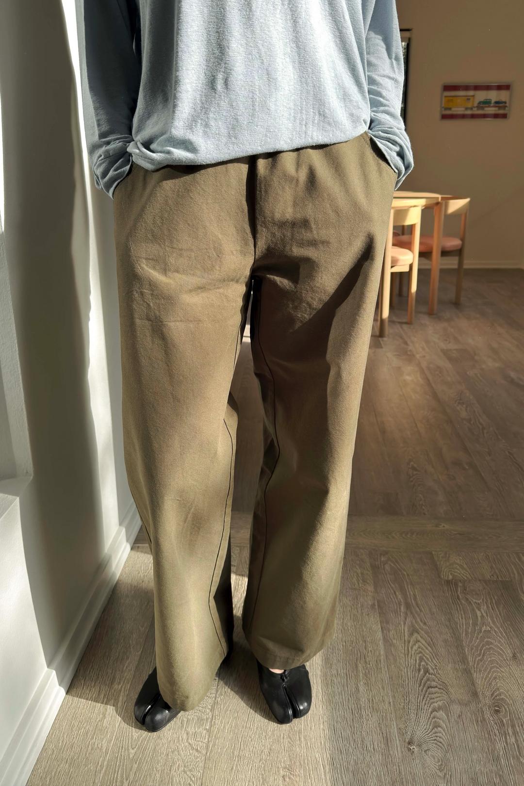 A person stands indoors with hands in pockets, wearing Le Bon Shoppe - Work Pant / Olive and a loose light gray long-sleeve shirt, paired with black split-toe shoes. Sunlight creates shadows on the wooden floor and nearby furniture.