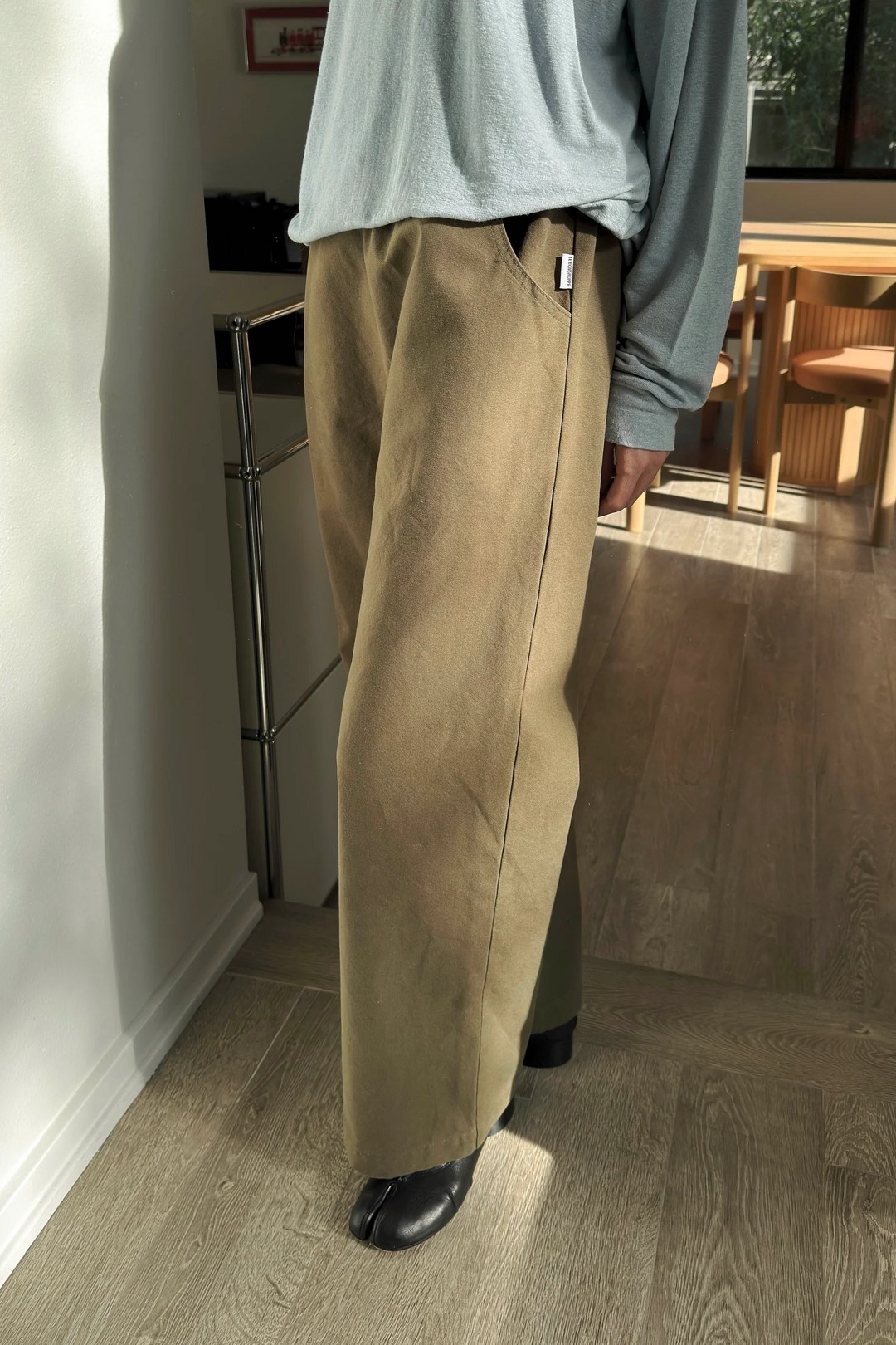 A person stands indoors on a wooden floor in natural sunlight, wearing Le Bon Shoppe - Work Pant / Olive and a light gray long-sleeved top with black shoes.