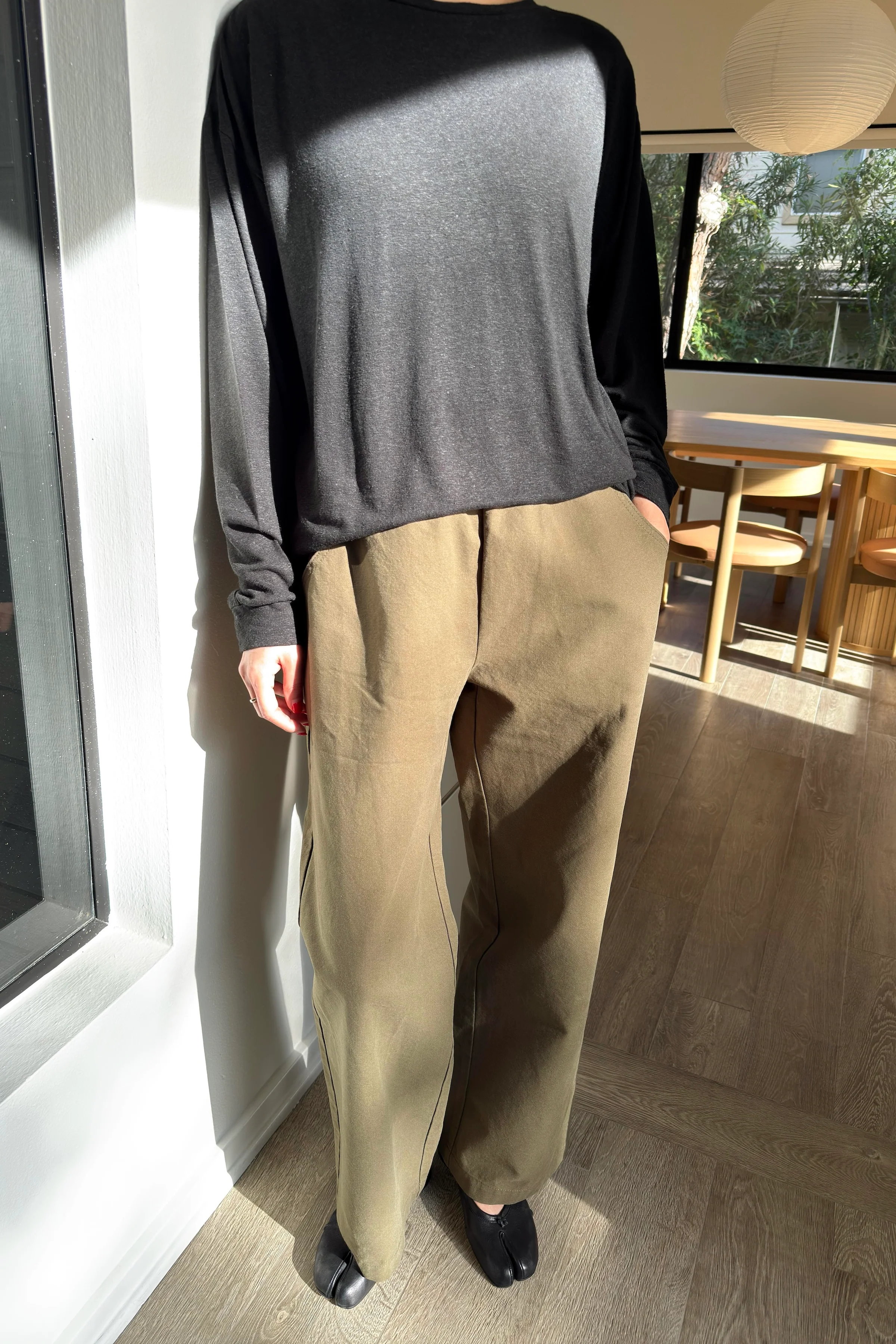 A person stands indoors by a window wearing a loose black long-sleeve top, black shoes, and Le Bon Shoppe - Work Pant / Olive by Le Bon Shoppe. Sunlight streams in, casting shadows on the wall and wooden floor.