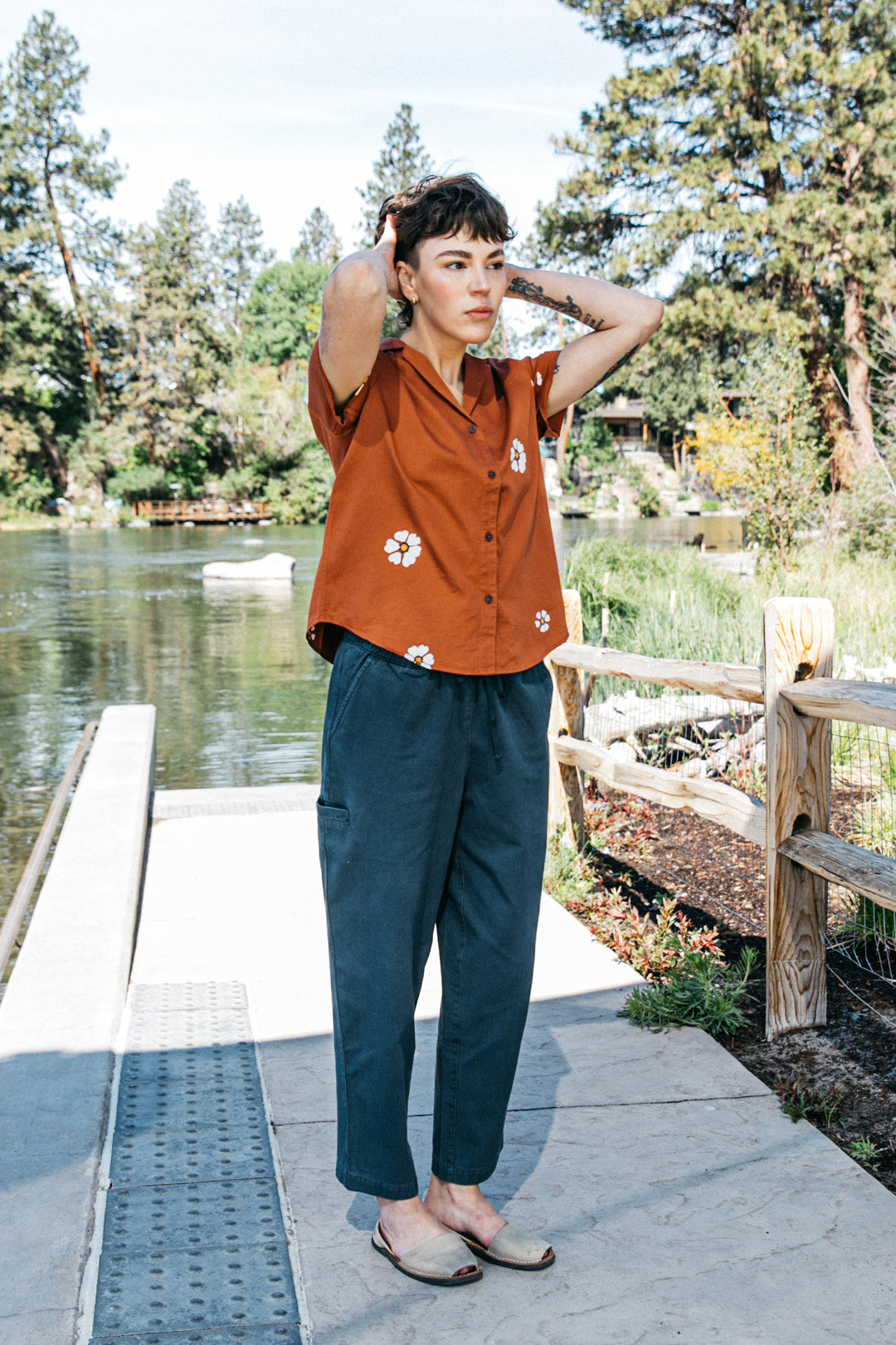 Outdoors by a river, a person wears the Bridge & Burn Wren Shirt in Redstone Bloom—featuring white flower prints and breathable cotton—paired with blue pants, enjoying comfort and nature's greenery.