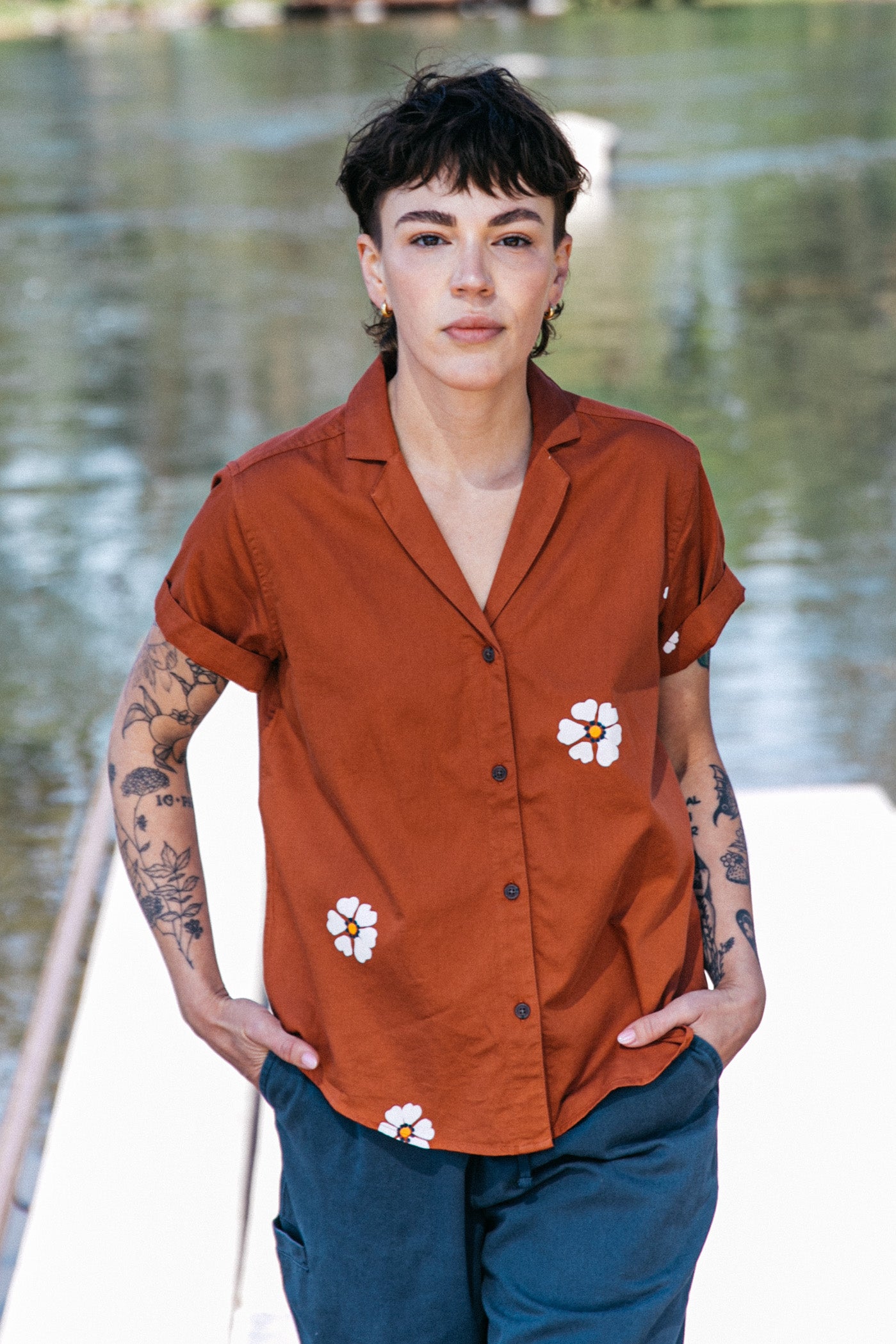 A person with short dark hair and tattoos stands outdoors by water, wearing the Bridge & Burn Wren Shirt in Redstone Bloom—a retro-inspired, rust-colored, breathable cotton shirt with white floral designs—paired with dark pants.
