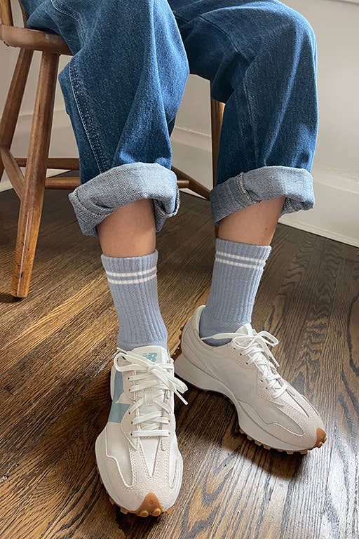 A person in cuffed blue jeans, Le Bon Shoppe - Boyfriend Socks in Blue Grey, and white sneakers sits on a wooden chair on a hardwood floor.