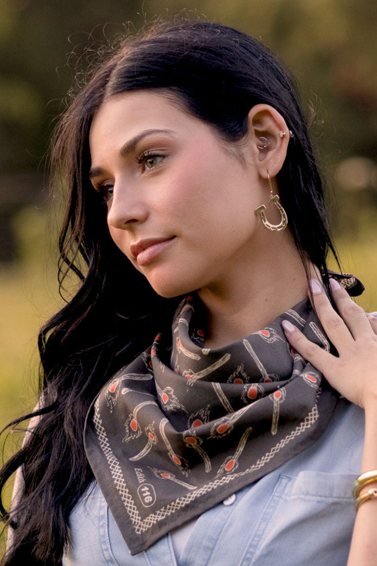 A woman with long dark hair, gold earrings, and a light blue shirt stands outdoors wearing the Handker Bandanas - No.116 Edith Bandana by Handker Bandanas around her neck, looking calmly to the side.