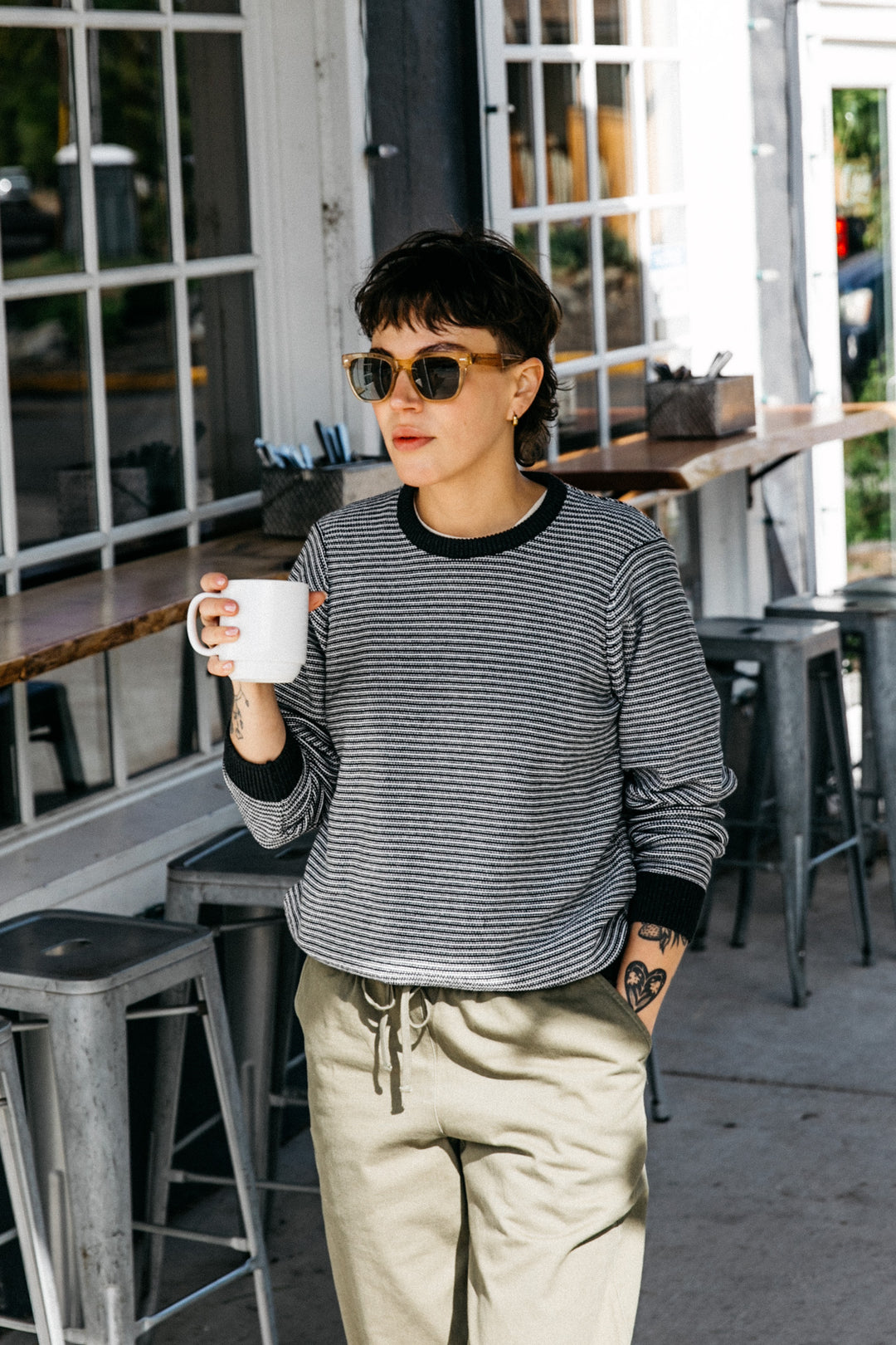 Wearing the Bridge & Burn Ansel Sweater in Charcoal Stripe, a person with short hair and tattoos stands outside a café, holding a white mug. They're in sunglasses, with high stools and large windows behind them.