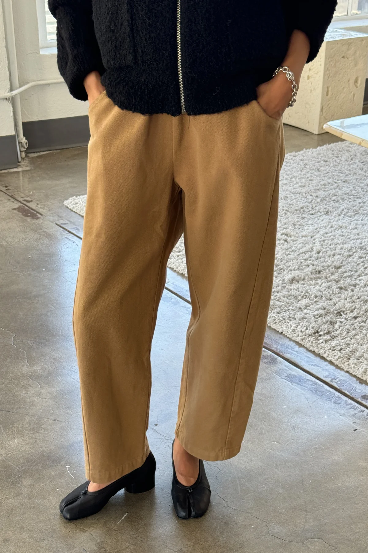Wearing Le Bon Shoppe’s Arc Pant in Tobacco, a person stands indoors on a concrete floor near a textured rug, hands in pockets, styled with a black zip-up jacket and black square-toed shoes.