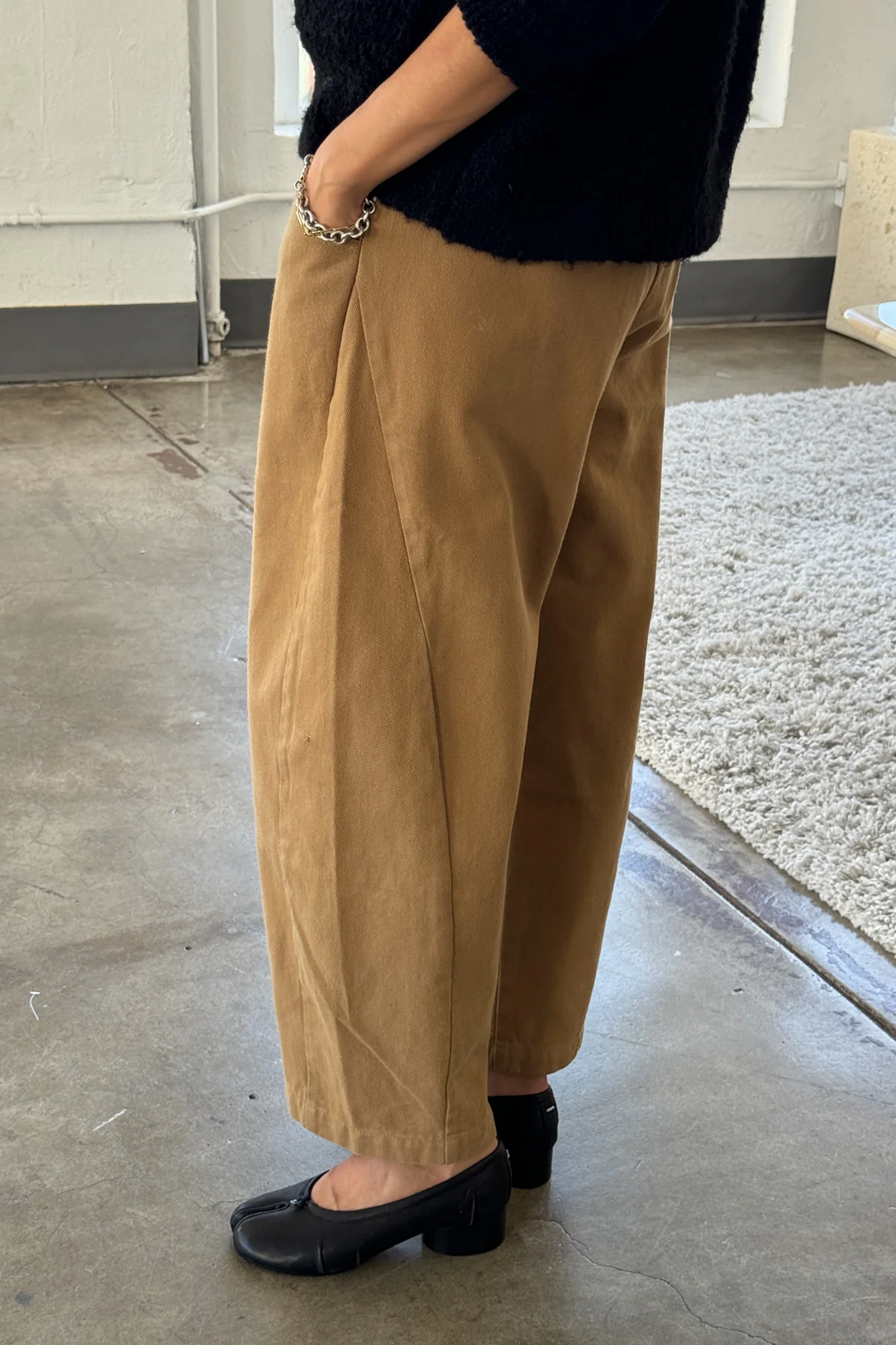 Indoors, a person (face not visible) wears loose Le Bon Shoppe Arc Pant in Tobacco, a dark sweater, black heeled shoes, and a silver bracelet. They're standing on a concrete floor with a textured cream rug.