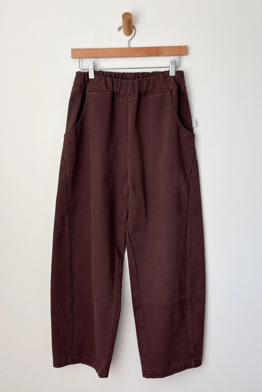 The Le Bon Shoppe - Arc Pant in Chocolate features a wide-leg, cropped fit with an elastic waistband and side pockets, crafted from heavyweight canvas. Shown on a wooden hanger against a plain white wall.