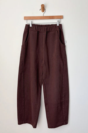 The Le Bon Shoppe - Arc Pant in Chocolate features a wide-leg, cropped fit with an elastic waistband and side pockets, crafted from heavyweight canvas. Shown on a wooden hanger against a plain white wall.