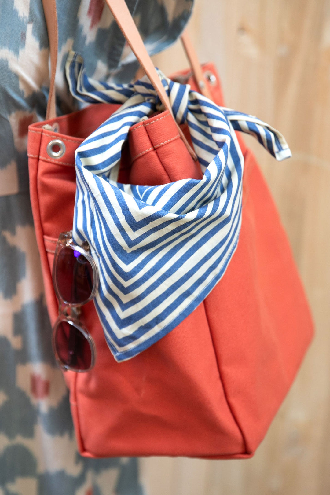 A close-up of an orange bag with a Handker Bandanas - No. 015 Sammie Bandana (22x22" cotton, hand screen-printed) tied to one handle and sunglasses hanging from the side, against a softly blurred background.