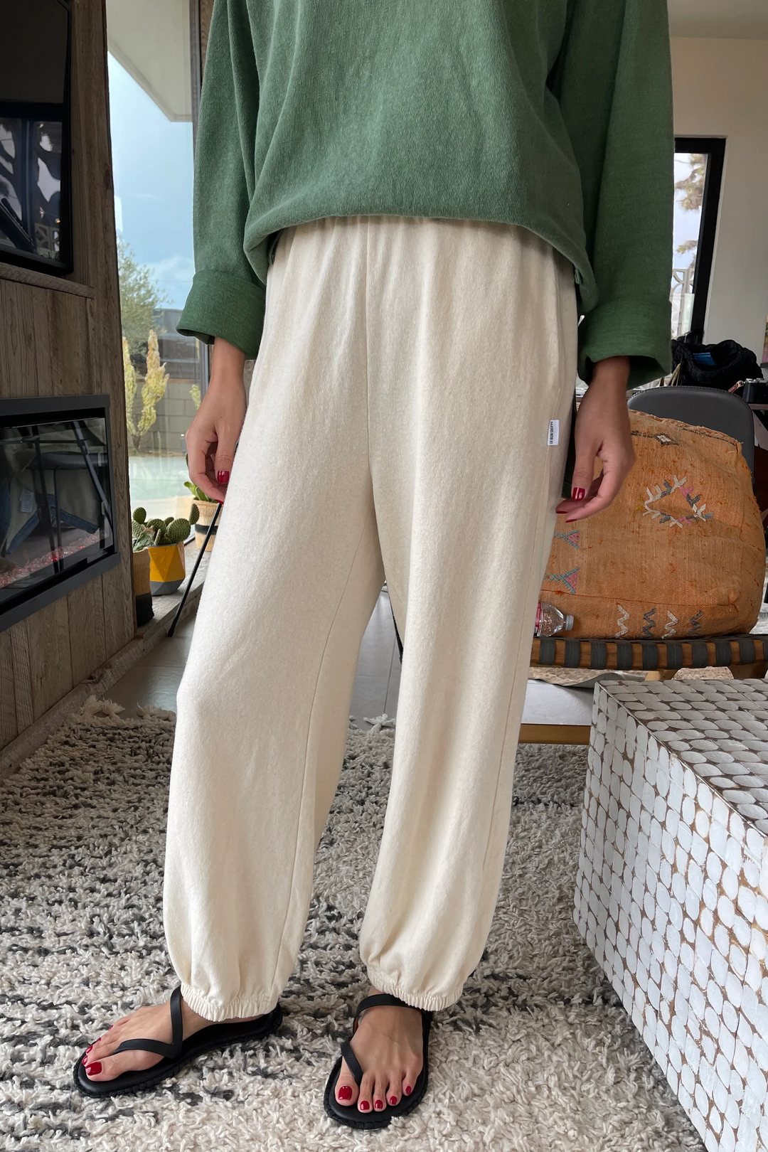 Only the lower body is visible as a person stands indoors on a textured rug, wearing Le Bon Shoppe - Balloon Pant in Natural and black sandals, with red-painted toenails.