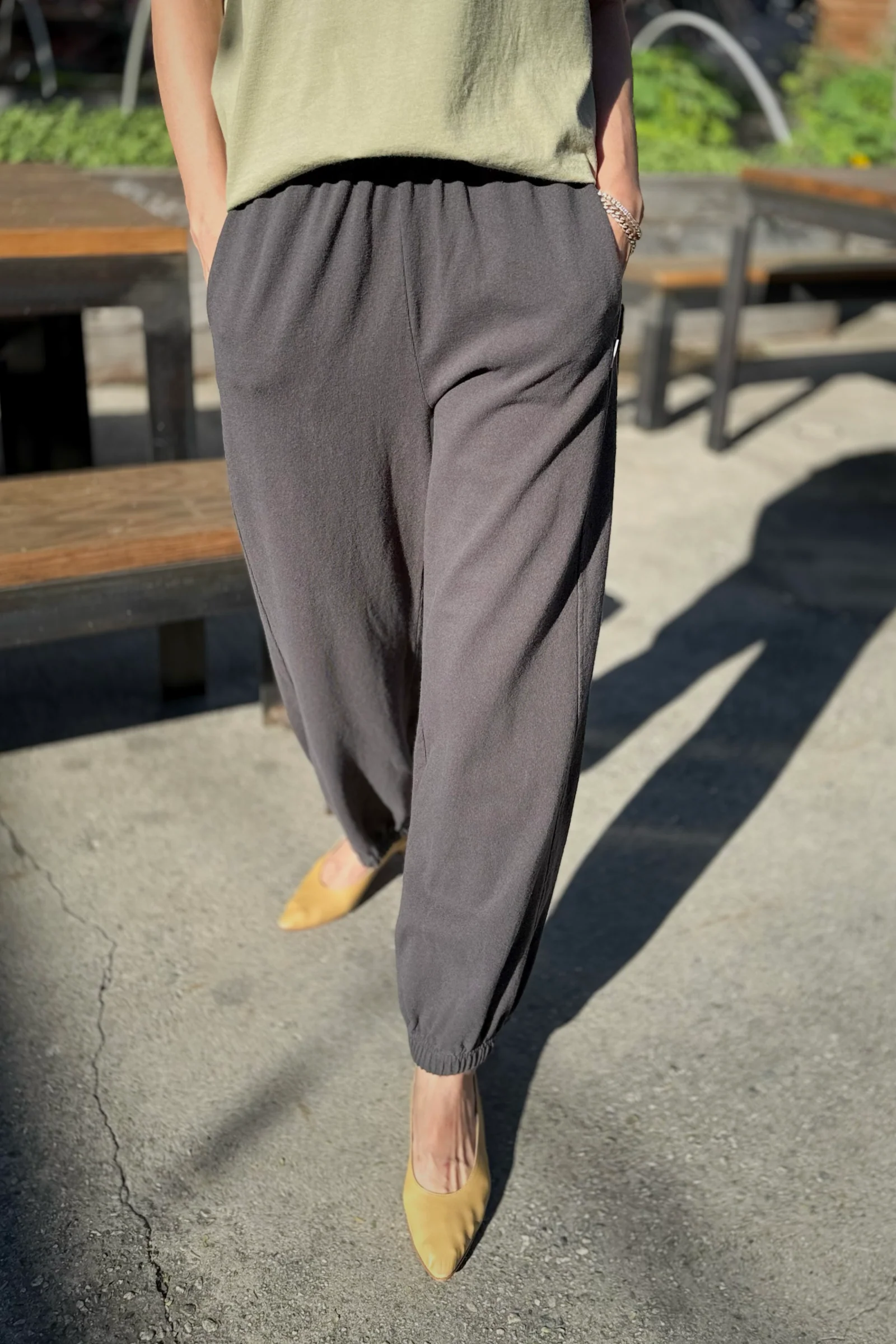 Wearing Le Bon Shoppe Balloon Pant in Dark Slate with yellow pointed-toe shoes, this person stands on a sunlit pavement, hands in pockets. Wooden benches and greenery form the background.