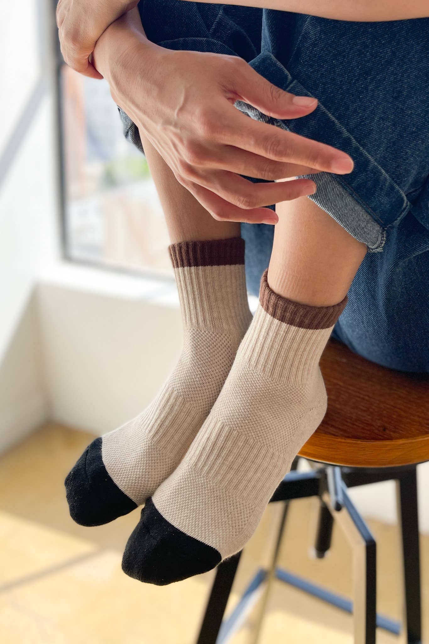 A person sits on a stool in rolled-up jeans and Le Bon Shoppe - Color Block Girlfriend Socks / Black Coffee, sunlight streaming in through the window.