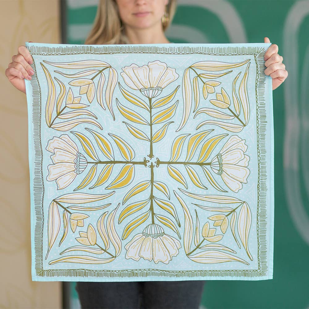 A person holds a square, light blue Handker Bandanas - No. 012 Eliza Bandana by Handker Bandanas, featuring white flowers, green and yellow leaves, and a striped border. The person's face is out of focus.
