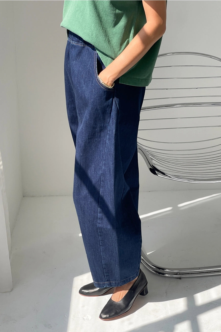 Indoors, a person poses with one hand in their pocket next to a modern metal chair, wearing Le Bon Shoppe - Arc Pant in Blue Denim. Sunlight casts shadows across the white floor.
