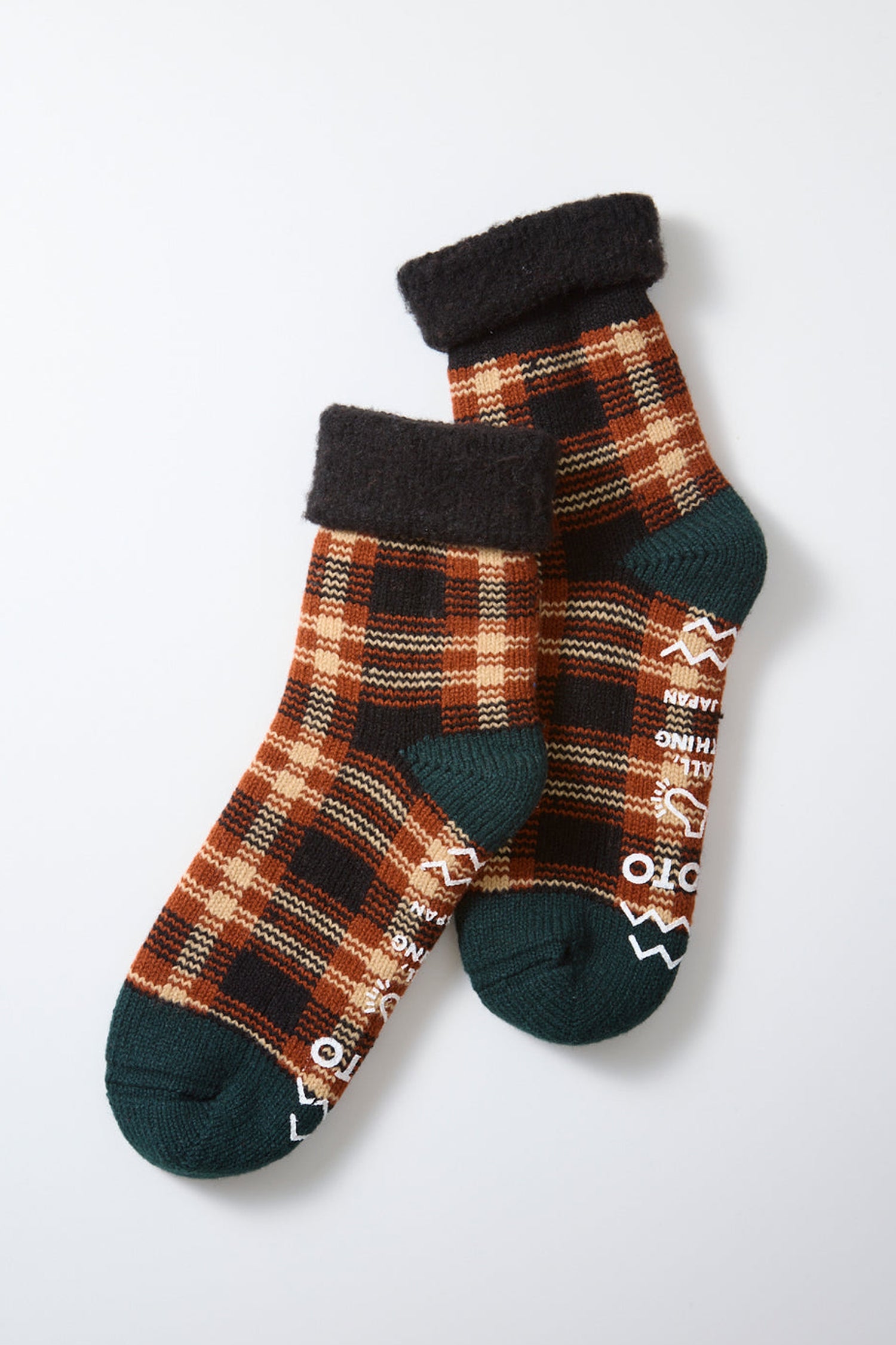 Two ankle-length RoToTo Comfy Room Check Socks in Black Brown, designed by RoToTo, showcase a brown, orange, and black plaid pattern with dark green toes and heels, complemented by plush black cuffs for added comfort. The soles feature white non-slip text and shapes to enhance grip on smooth surfaces. Made from a warm wool blend for softness and insulation, the socks are displayed overlapping on a white background.