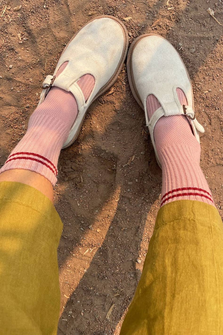 A person wearing mustard yellow pants, Le Bon Shoppe’s Boyfriend Socks in Vintage Pink, and light beige Mary Jane shoes stands on sunlit dirt.