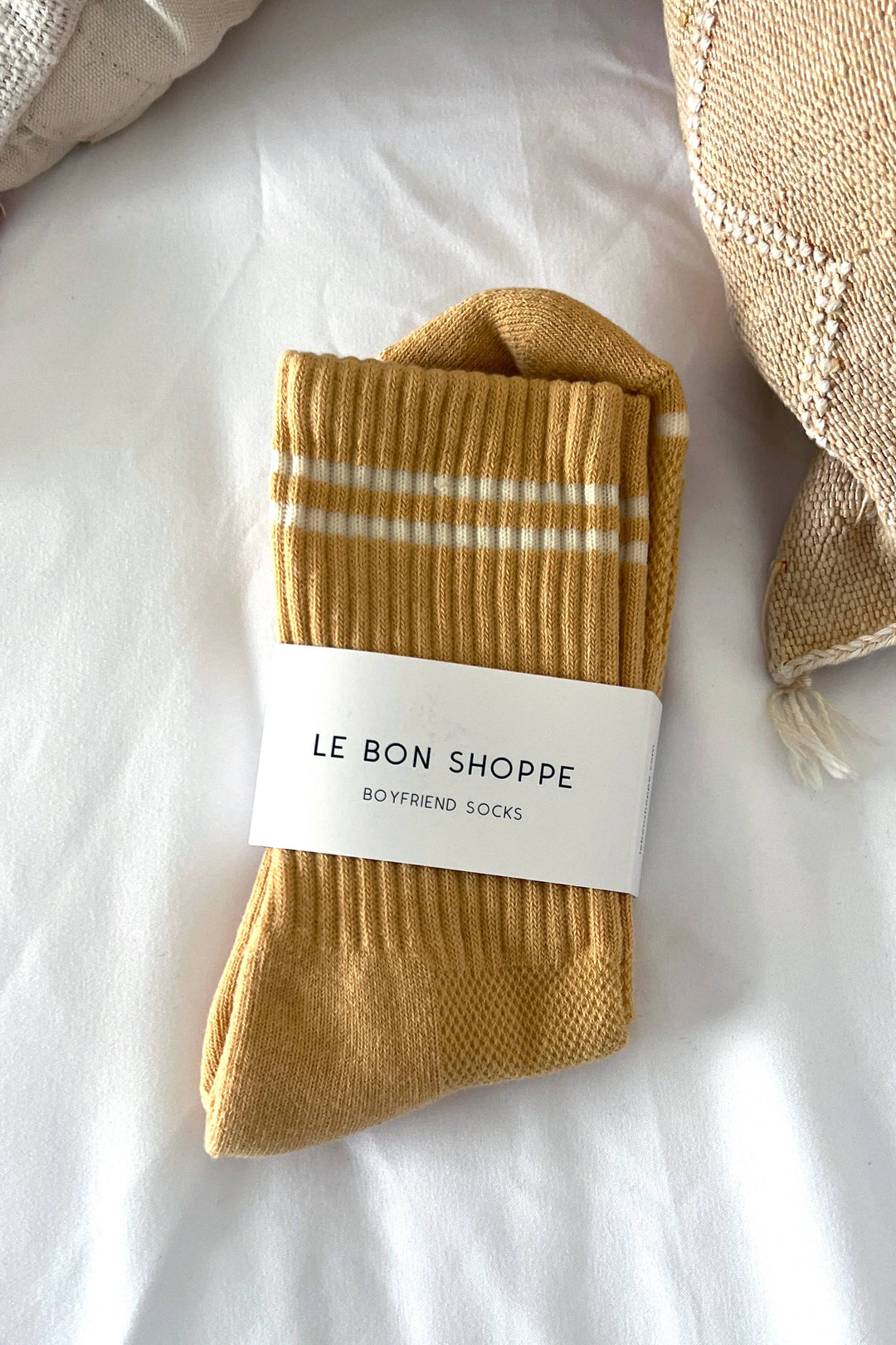 A pair of Le Bon Shoppe Boyfriend Socks in Butter, with white stripes and the brand’s label, is neatly folded on a white surface next to beige textured cushions.