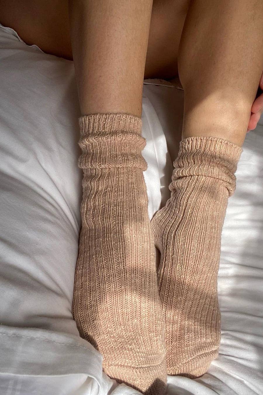 A close-up of a person's lower legs and feet wearing Le Bon Shoppe - Cottage Socks in Peachy Keen, resting on a white bed with sunlight casting soft shadows.