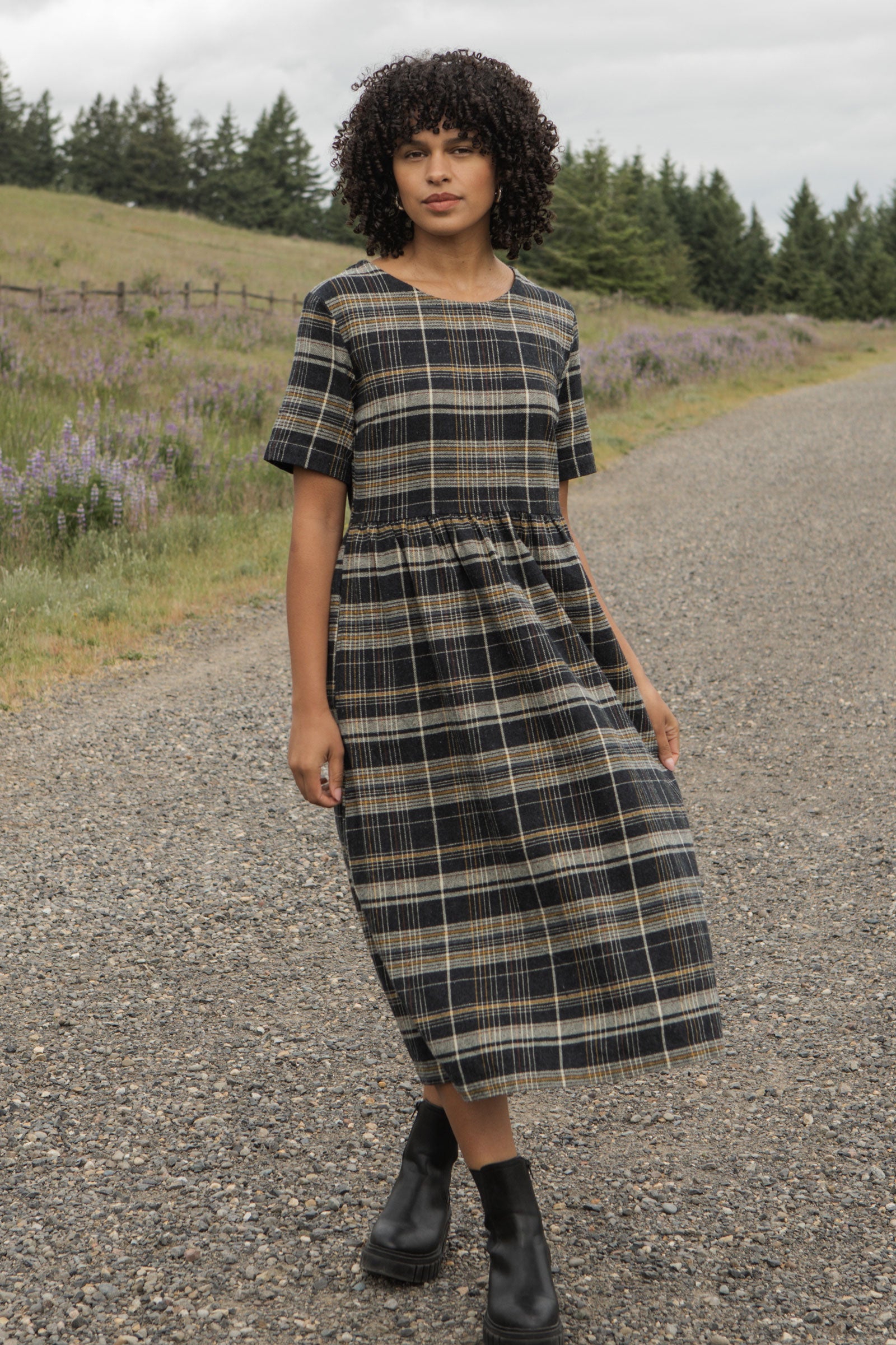 The Perry Dress in Volcanic Plaid by Bridge & Burn is a midi dress with short sleeves, made from recycled cotton. It features a dark navy, yellow, and white plaid pattern with a fitted bodice and gathered skirt.