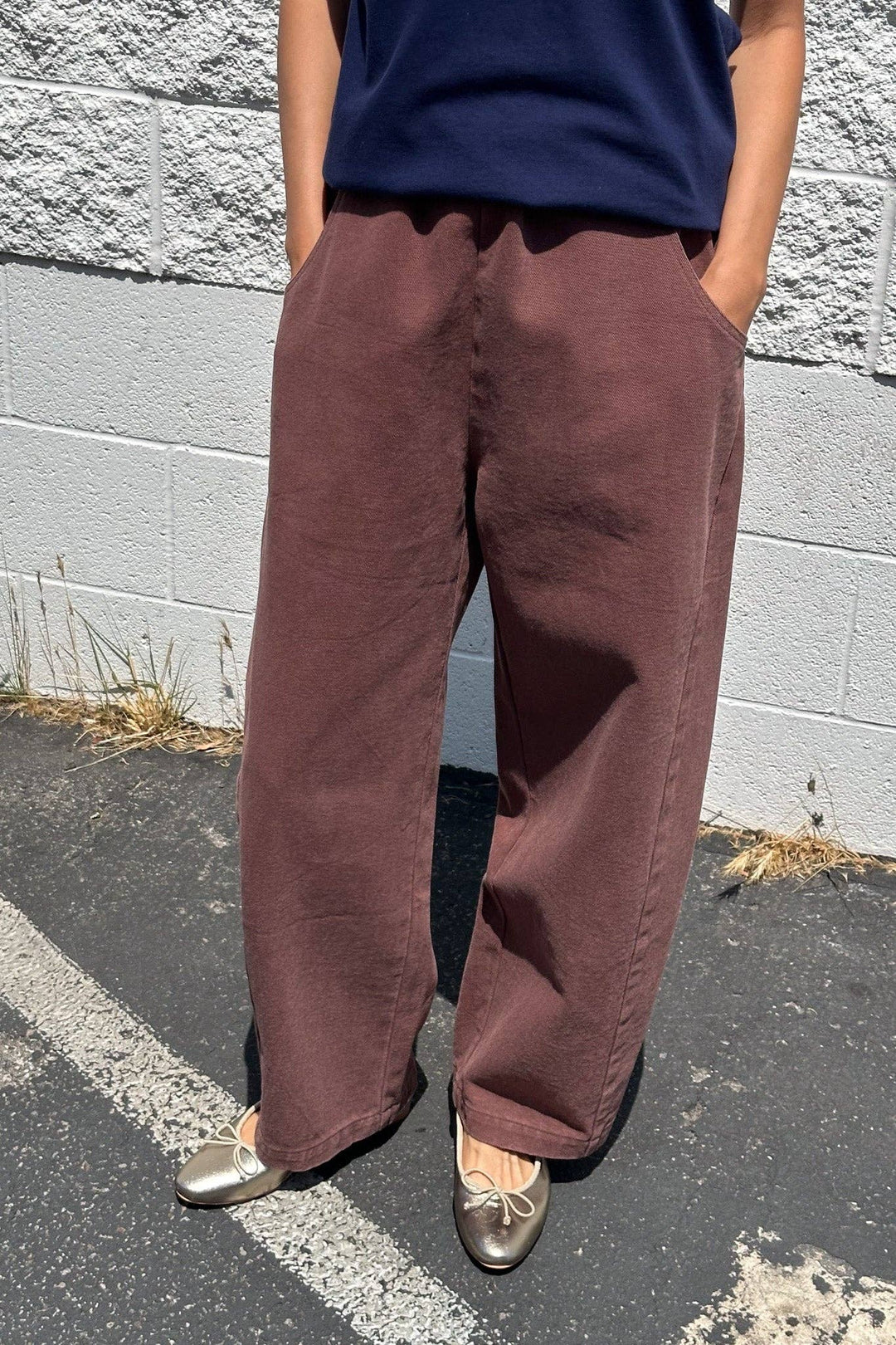 Someone stands outdoors on pavement, hands in the pockets of loose, high-waisted Le Bon Shoppe - Arc Pant in Chocolate, styled with a navy blue shirt and metallic silver ballet flats. A white brick wall and dry grass are in the background.