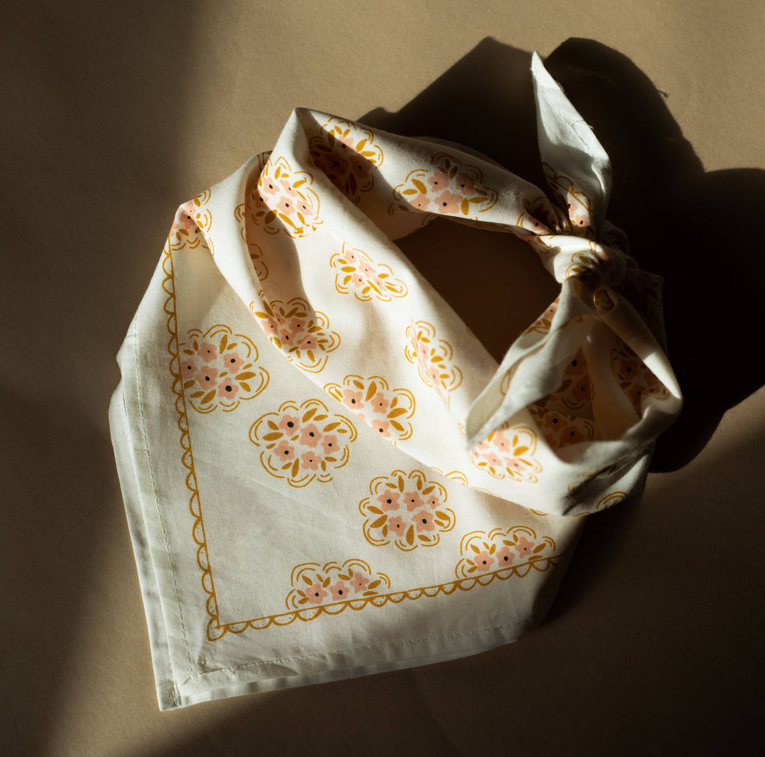 The Handker Bandanas - No. 042 Pearl Bandana by Handker Bandanas, featuring a cream cotton fabric with yellow floral motifs and a scalloped border, is folded on a brown surface with soft natural sunlight and gentle shadows.