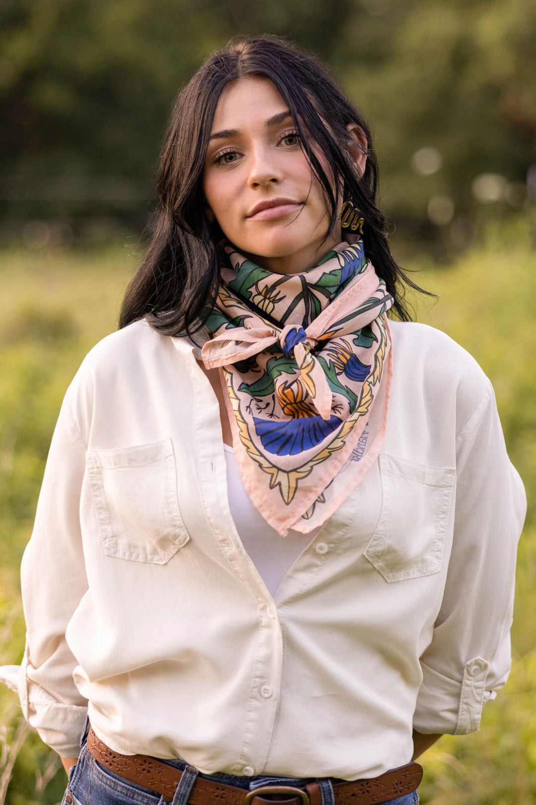 A woman with long dark hair stands outdoors, wearing a cream cotton button-up shirt and the Handker Bandanas 34" Dandy Wild Rag Bandana as a colorful floral scarf. A blurred green field with trees is in the background.