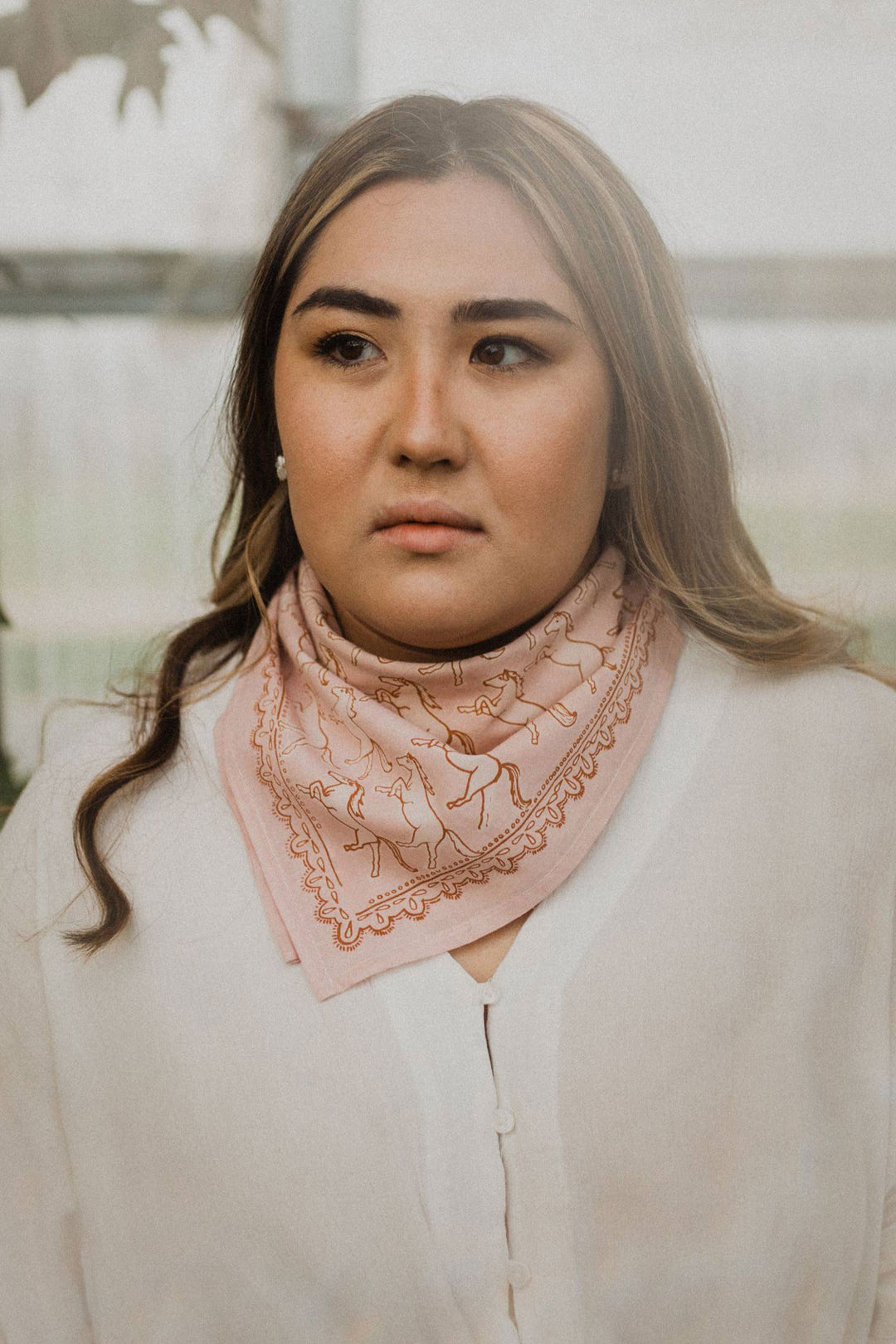 A woman with long brown hair wears a white blouse and the Handker Bandanas No. 062 Horses Bandana, gazing slightly to the side with a neutral expression against a softly blurred background.