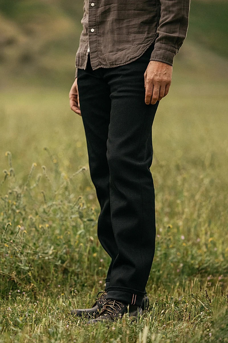 A person stands in a grassy field, wearing a brown button-up shirt and Hiroshi Kato - The Hammer Straight / Black Raw 14oz Selvedge jeans. Their dark shoes complete the look, with the blurred background highlighting their lower body and legs.