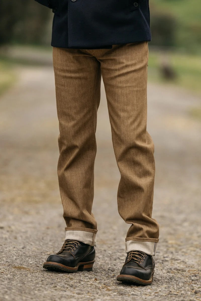A person stands on a gravel path wearing Hiroshi Kato - The Hammer Straight / Copper Raw 10.5oz Selvedge jeans, the cuffs rolled up to reveal the Japanese selvedge denim, paired with black lace-up shoes featuring brown soles. Only their lower half is visible, with a backdrop of blurred greenery.