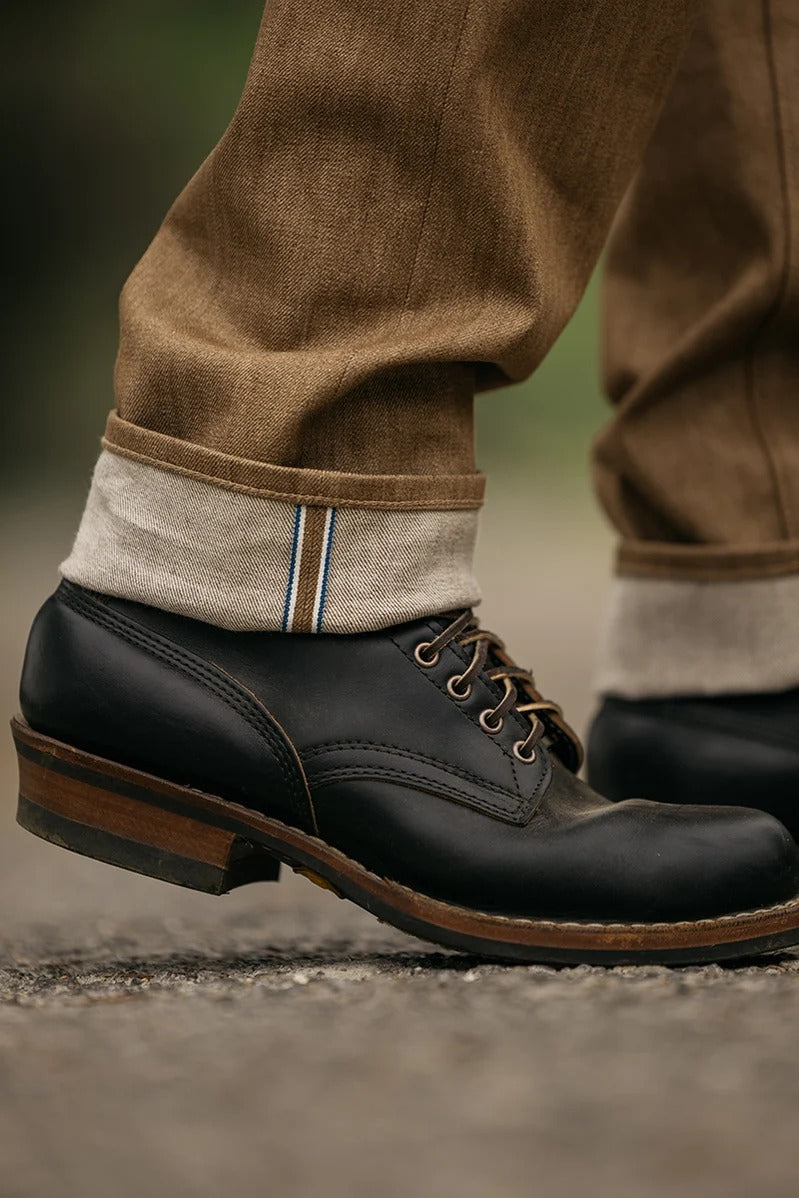 Close-up of a person wearing Hiroshi Kato - The Hammer Straight / Copper Raw 10.5oz Selvedge jeans, cuffed to reveal the Japanese selvedge edge, paired with black leather dress shoes and standing on a textured outdoor surface.