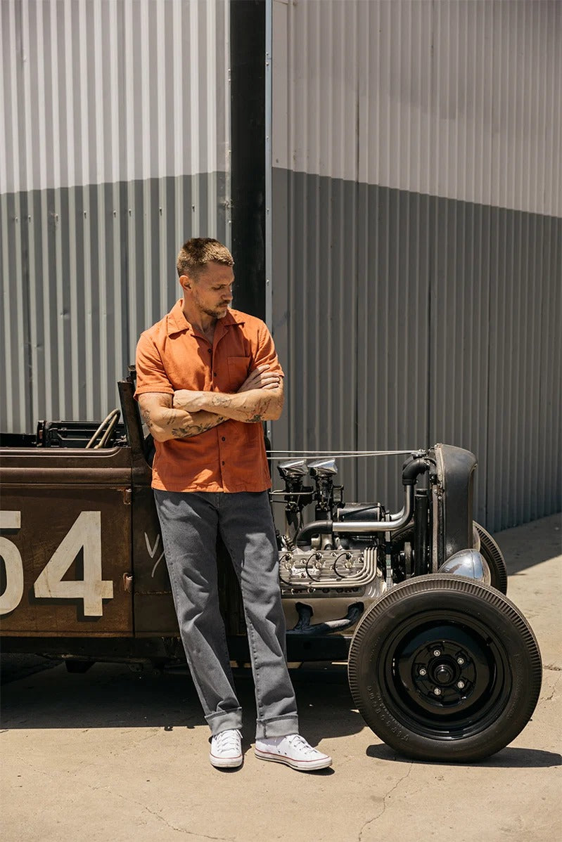 A man in an orange shirt and gray pants, likely Hiroshi Kato - The Hammer Straight / Granite 10.5oz Selvedge from Hiroshi Kato, stands with arms crossed beside a vintage car with the number 54, parked outside a corrugated metal building.