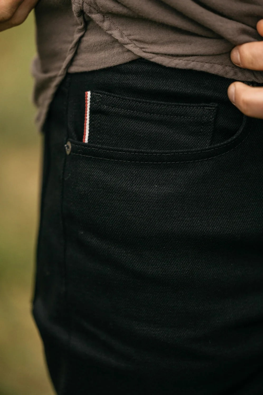 Close-up of a person wearing the Hiroshi Kato - The Hammer Straight / Black Raw 14oz Selvedge jeans, crafted from premium Japanese denim with a distinctive red and white striped selvedge detail on the pocket. The individual has a brownish shirt tucked in, and their hand rests near the waistband.