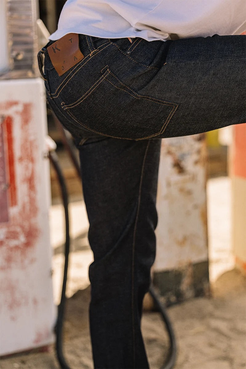 A person wearing Hiroshi Kato - The Hammer Straight / Indigo Raw 14oz Selvedge jeans stands near an old gas pump with one leg bent, the leather patch on the back pocket from Hiroshi Kato clearly visible.