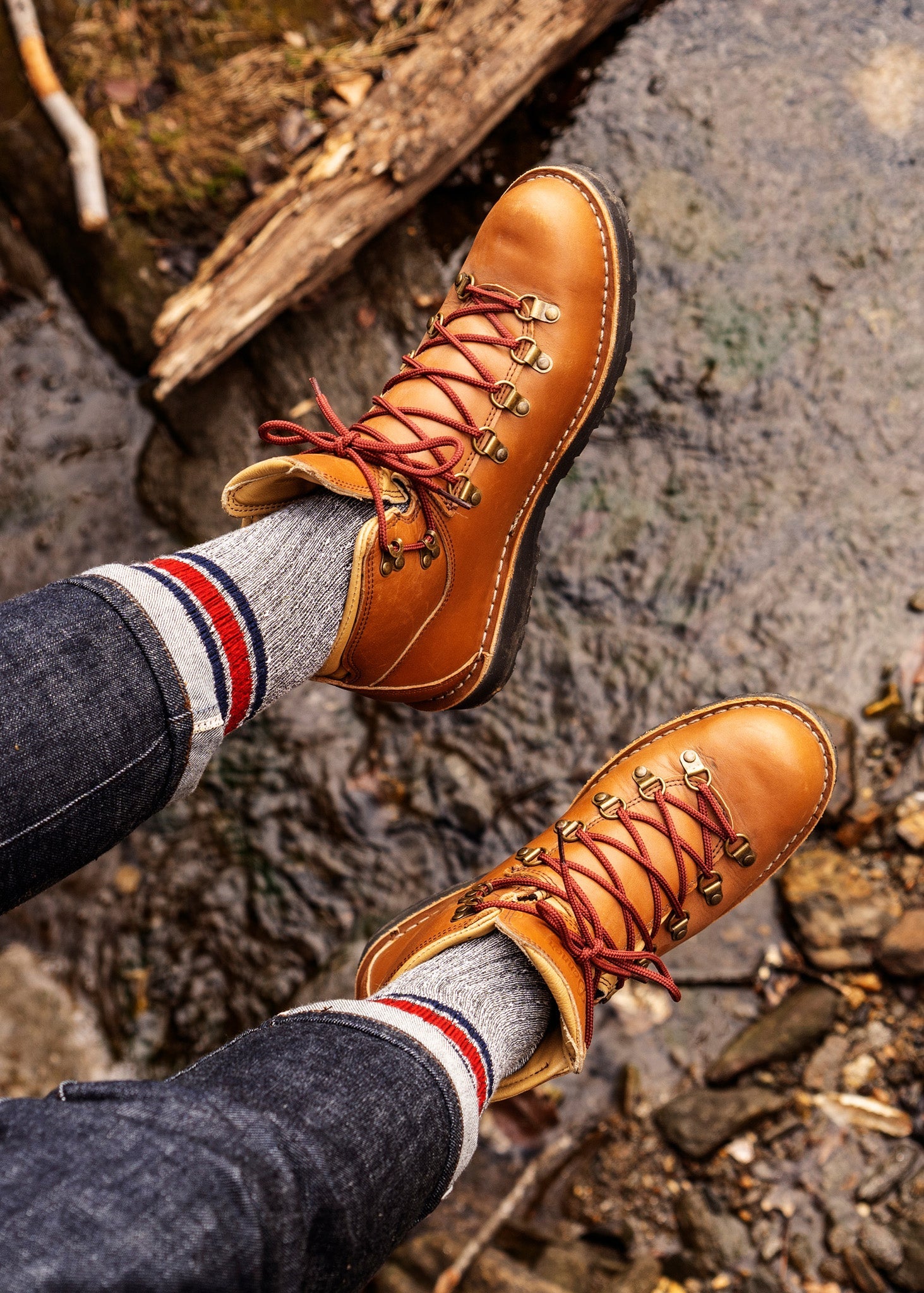 A person wearing American Trench Merino Activity Crew socks in grey, brown leather hiking boots, and dark jeans stands on rocky ground by water and wooden debris.