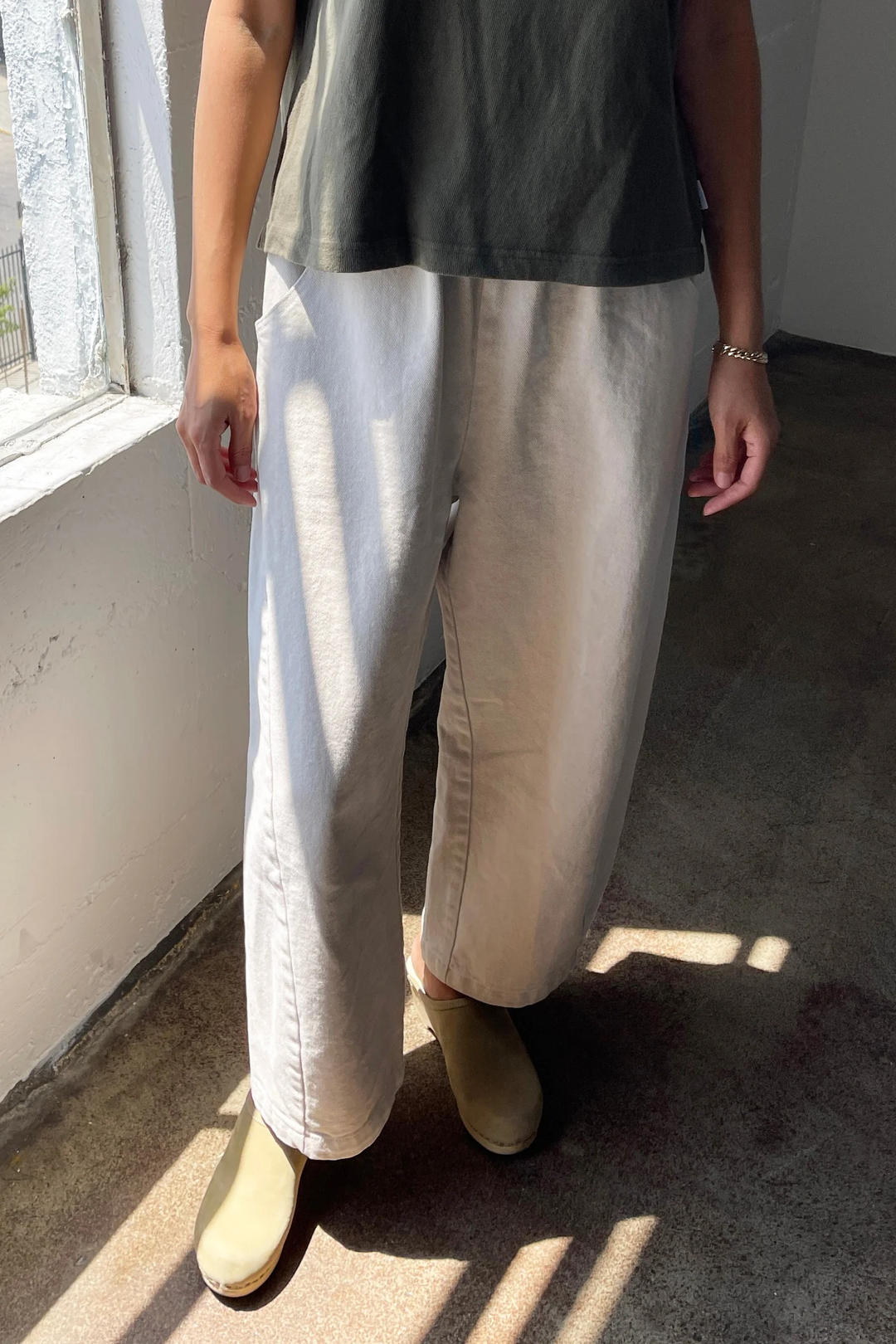 A person stands indoors by a sunlit window, wearing a dark short-sleeve shirt, Le Bon Shoppe - Arc Pant in Natural, olive green shoes, and a bracelet. Sunlight casts shadows on their clothes and the floor.