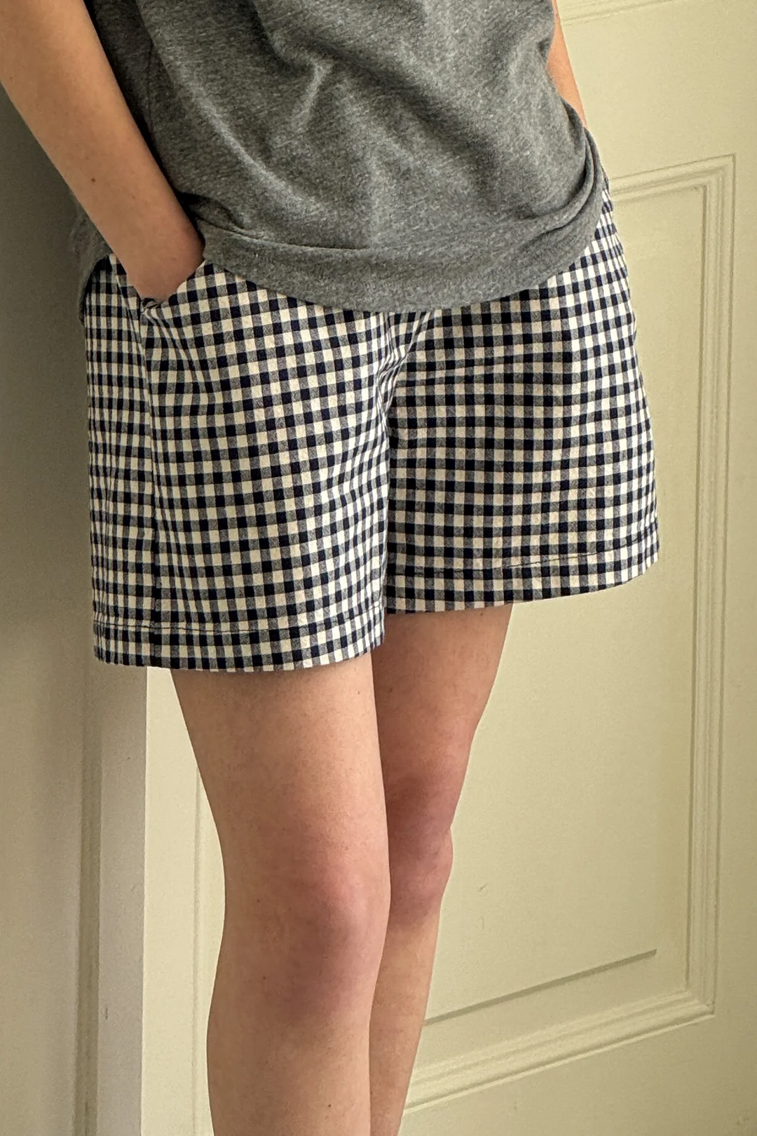 A person stands with hands in pockets, wearing a gray tee and Le Bon Shoppe - City Short / Navy Gingham. Only their torso and legs are visible in front of a light-colored door.
