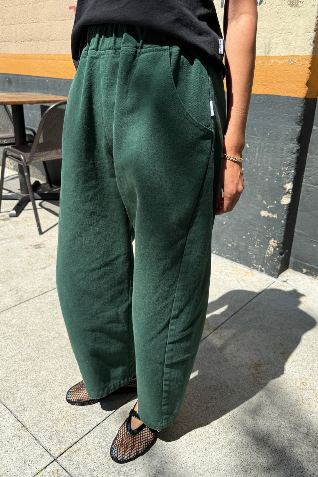 A person stands outdoors wearing Le Bon Shoppe - Arc Pant / Pine, paired with a black shirt and black mesh ballet flats. Sunlight casts shadows as only their lower body is visible on the concrete sidewalk.
