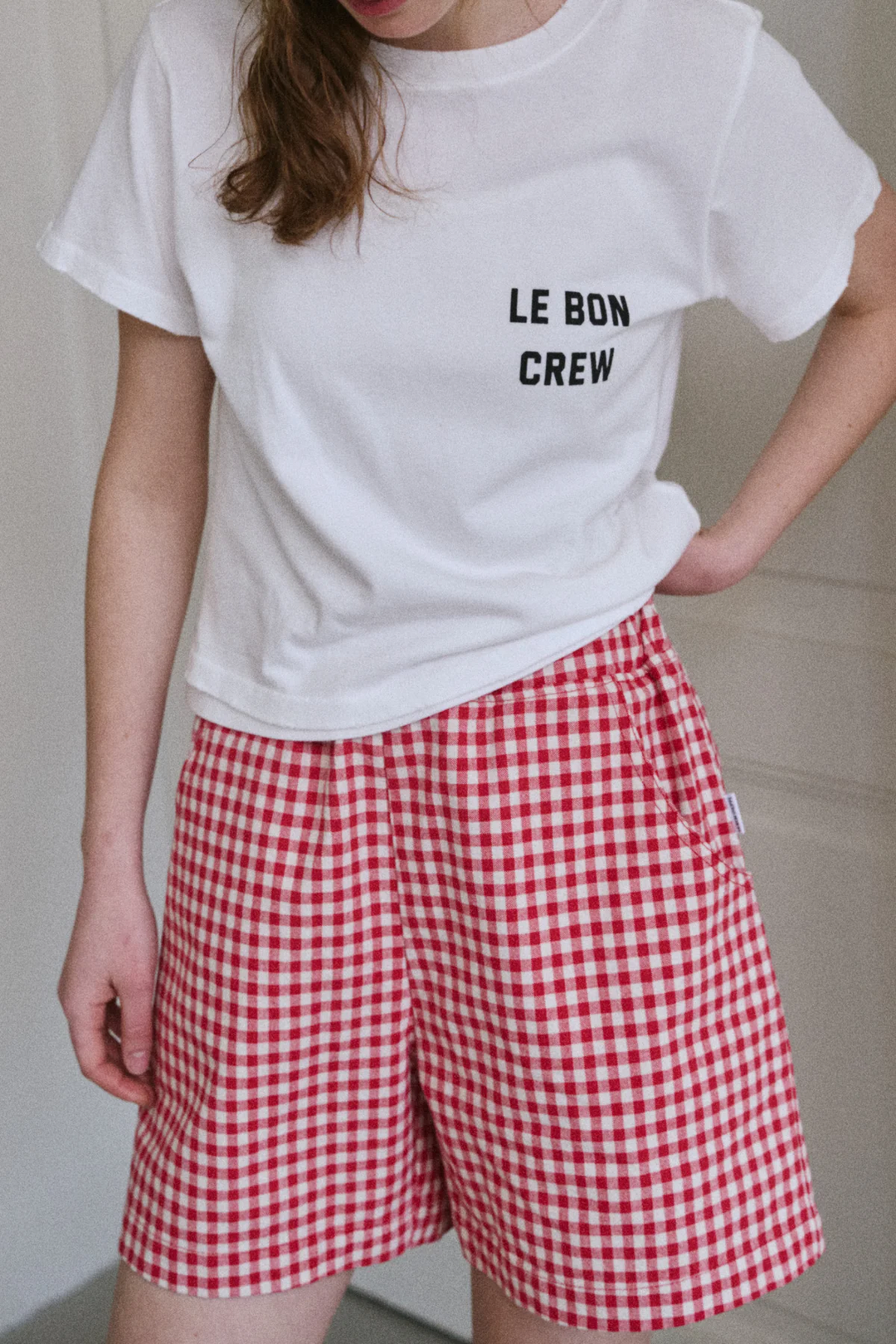A person stands indoors with one hand on their hip, wearing a white LE BON CREW t-shirt and Le Bon Shoppe - City Short in Red Gingham. Their face is partially out of frame.