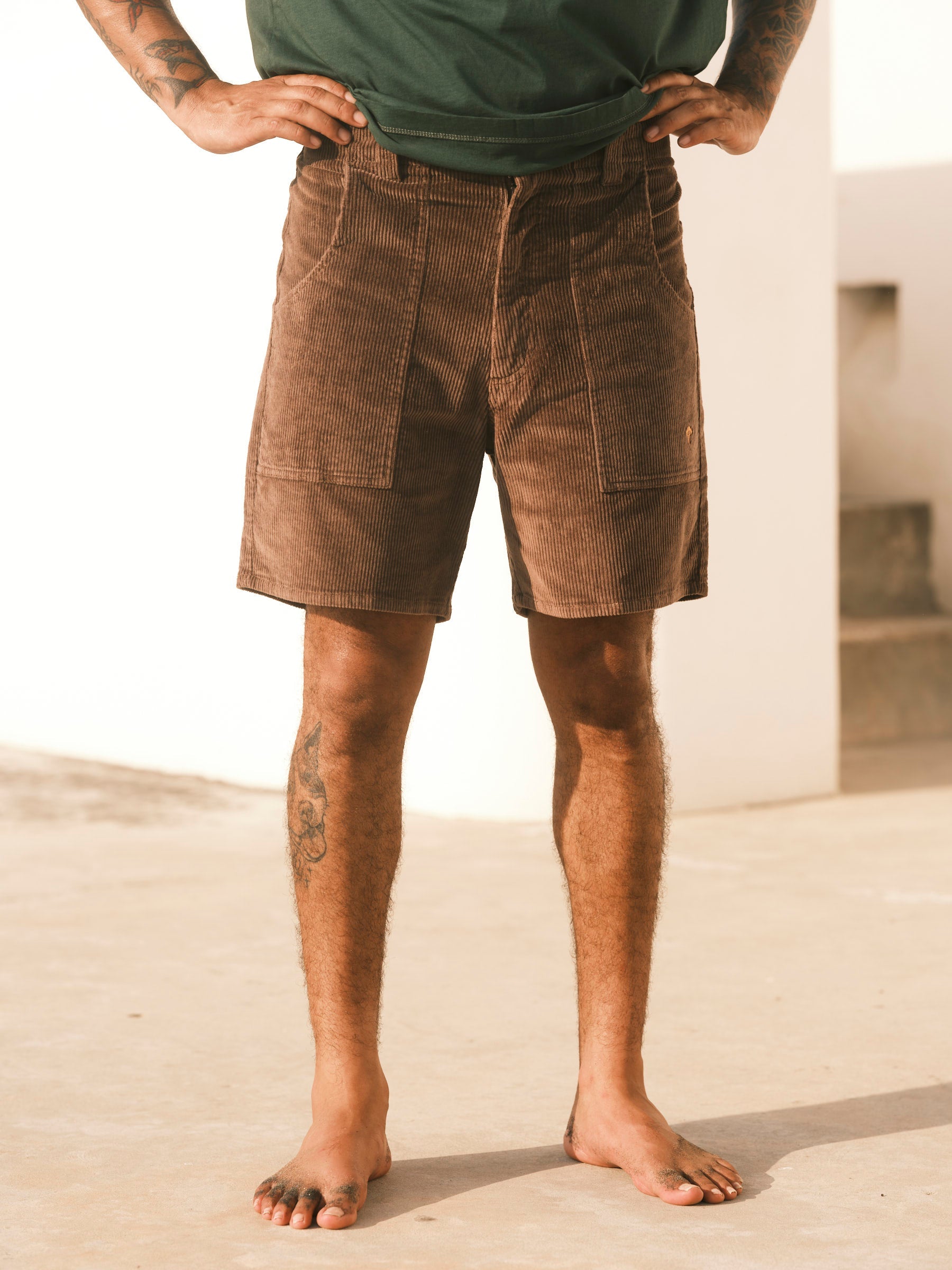 A person stands barefoot on concrete, hands on hips, wearing Mollusk Men's Shroom Shorts in Chocolate and a green shirt. They are dressed comfortably for warm weather. Their head is not shown in the image.