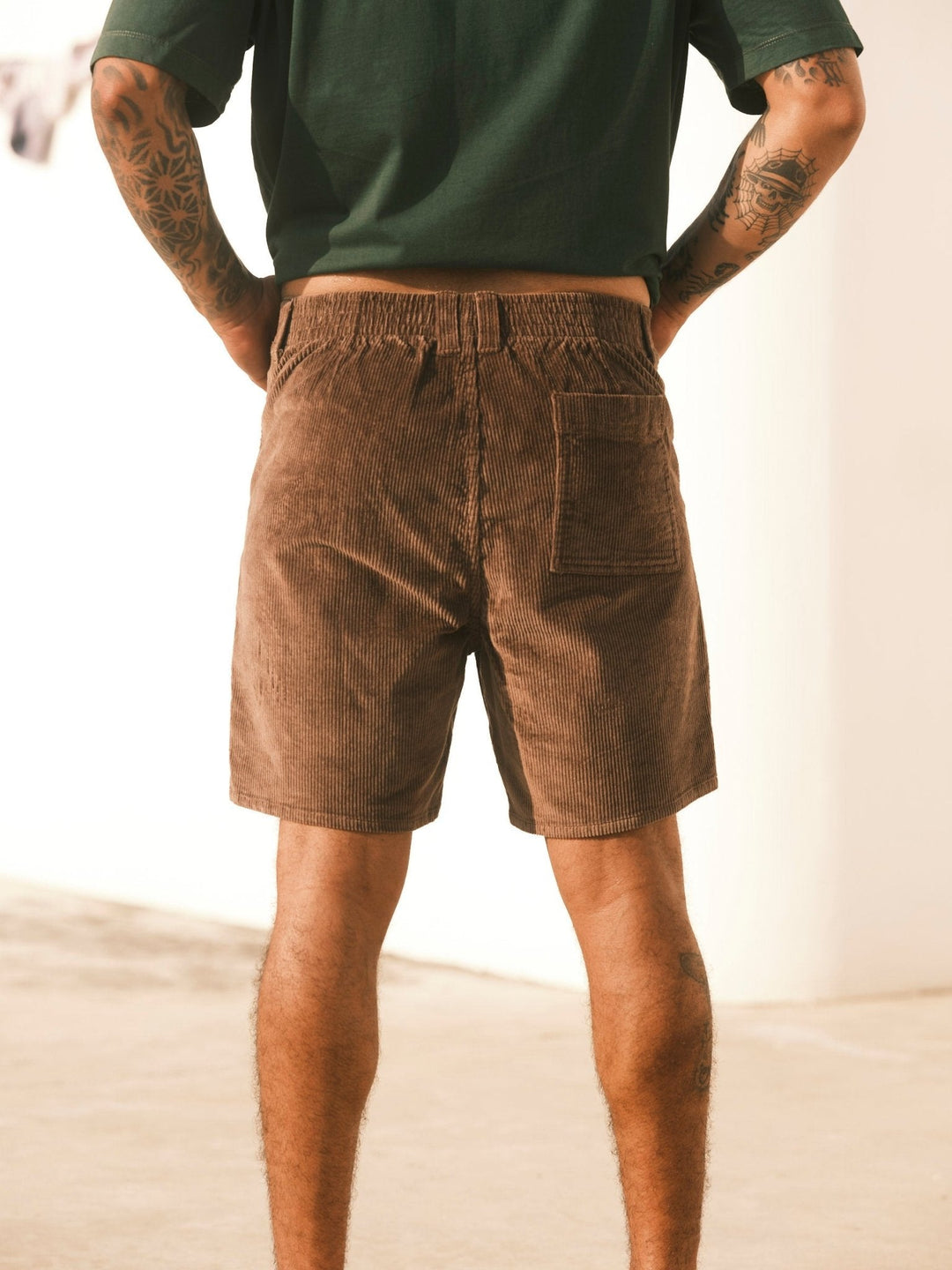 Indoors, a person stands with hands on hips in a dark green shirt and Mollusk - Men's Shroom Short / Chocolate by Mollusk. The photo from behind shows their tattooed arms and legs—ideal shorts for warm weather.